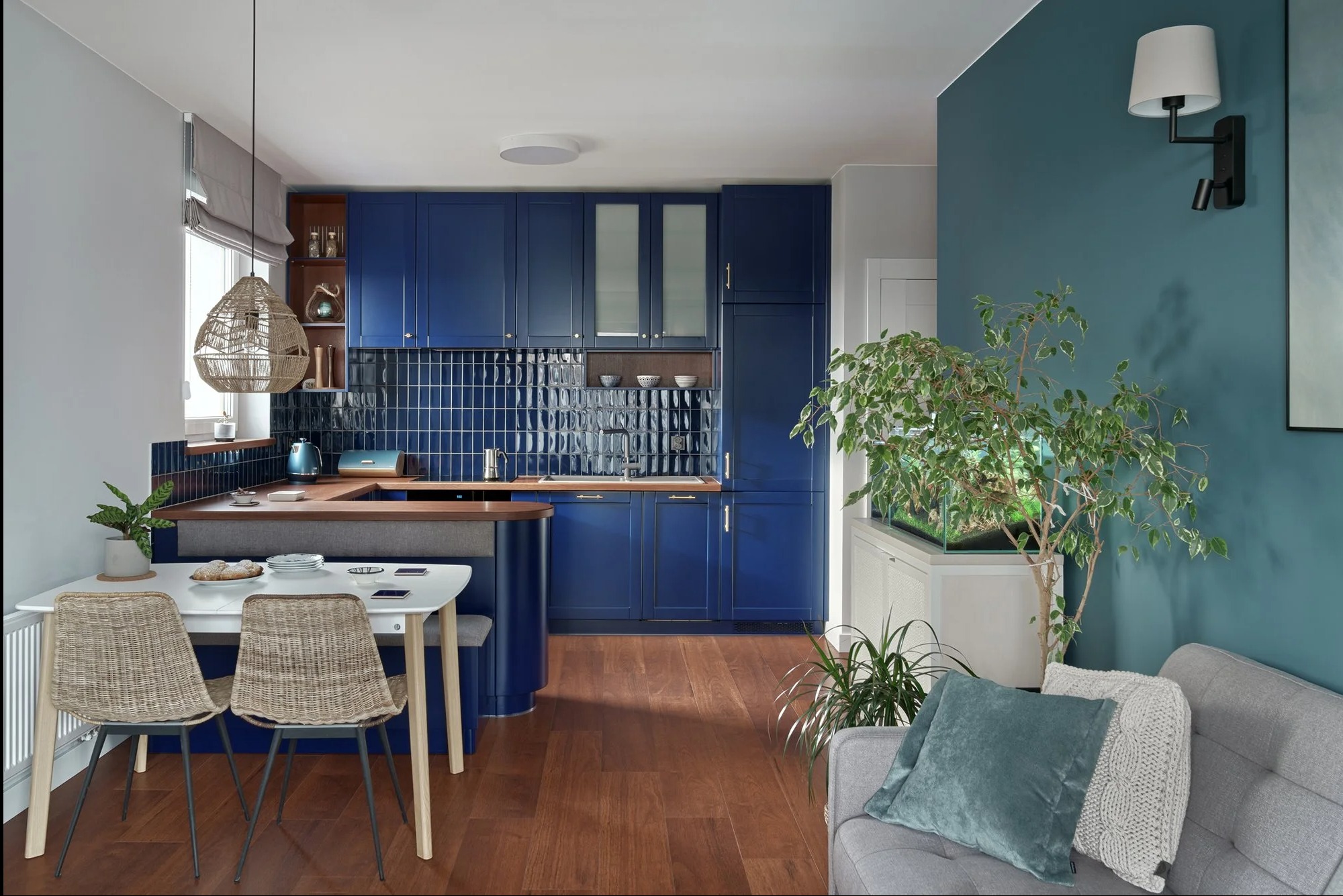 Nowoczesna kuchnia z niebieskimi szafkami, ciemnymi płytkami na ścianie, drewnianym blatem i podłogą. Stół z plecionymi krzesłami i lampą z naturalnych materiałów. W tle salon z szarą sofą...