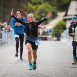 Uśmiechnięta biegaczka z numerem 131 na mecie biegu ulicznego, w tle kibice i kamerzysta.