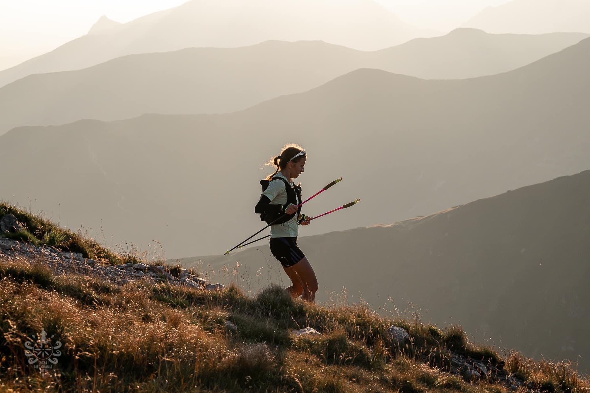 Kobieta biegnie w górach z kijkami trekkingowymi na tle zalesionych wzgórz w porannym świetle.