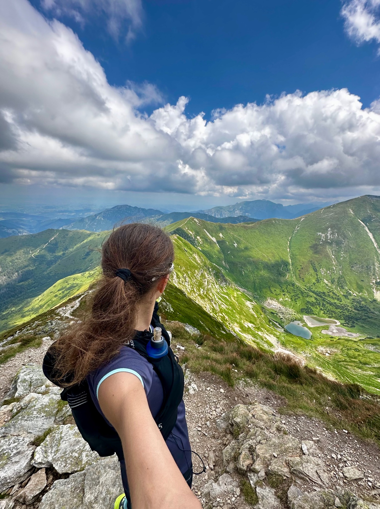 Kobieta z plecakiem biegowym spogląda na krajobraz górski z widocznymi szczytami, jeziorami i malowniczymi chmurami na niebie.