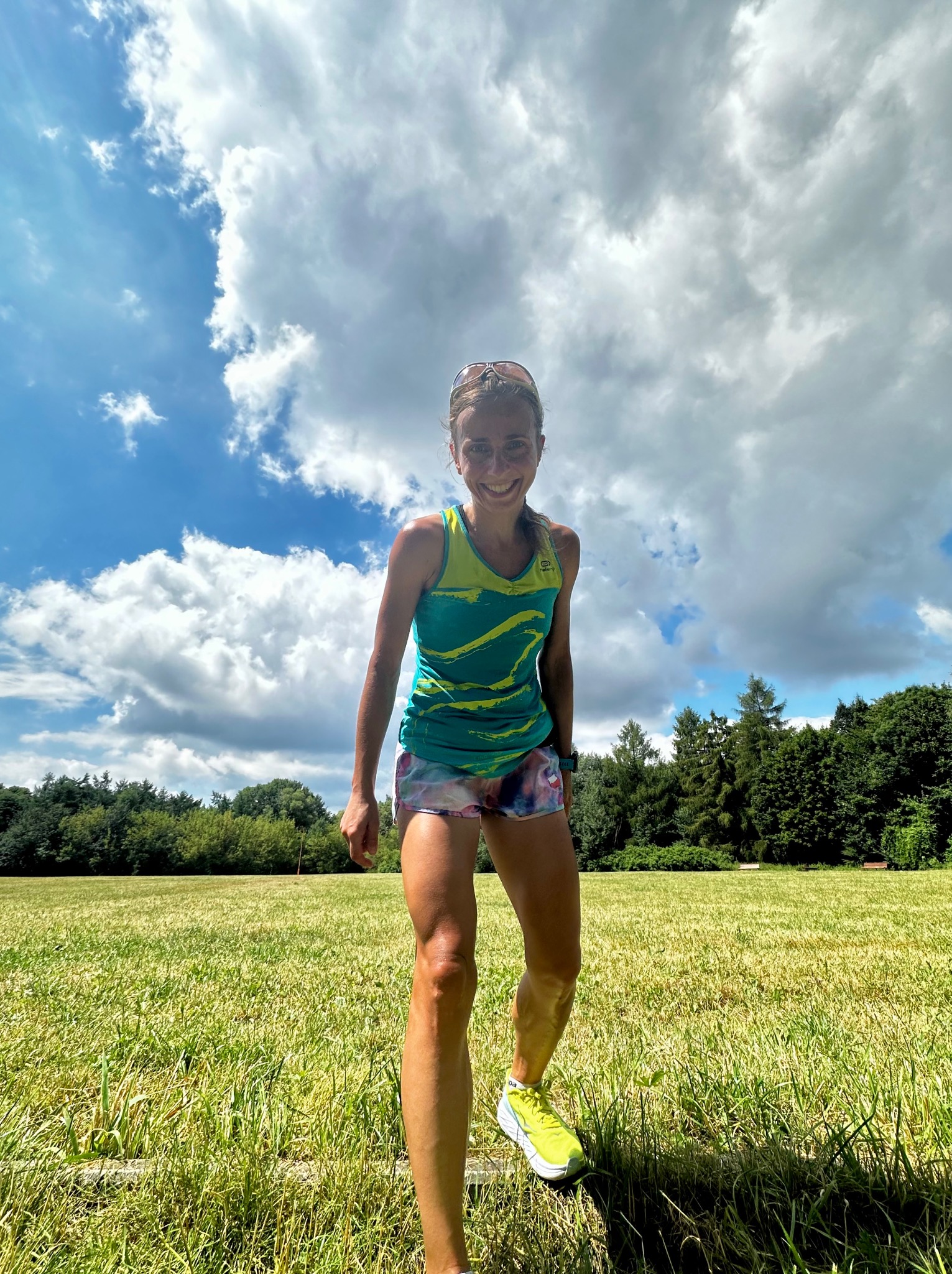 Uśmiechnięta kobieta w sportowym stroju, na zielonym polu z lasem w tle, pozuje w słoneczny dzień pod błękitnym niebem z chmurami, widoczny zegarek sportowy na nadgarstku.