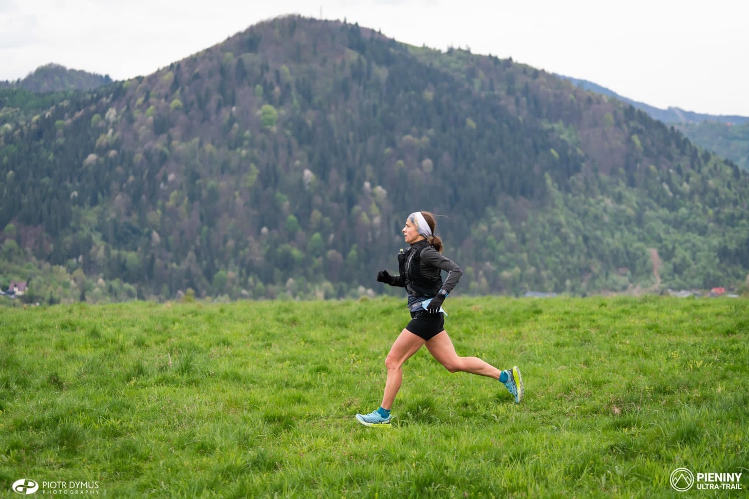 Kobieta w sportowym stroju biegnie przez zieloną łąkę, w tle zalesione wzgórze i logo Pieniny Ultra-Trail na dole.