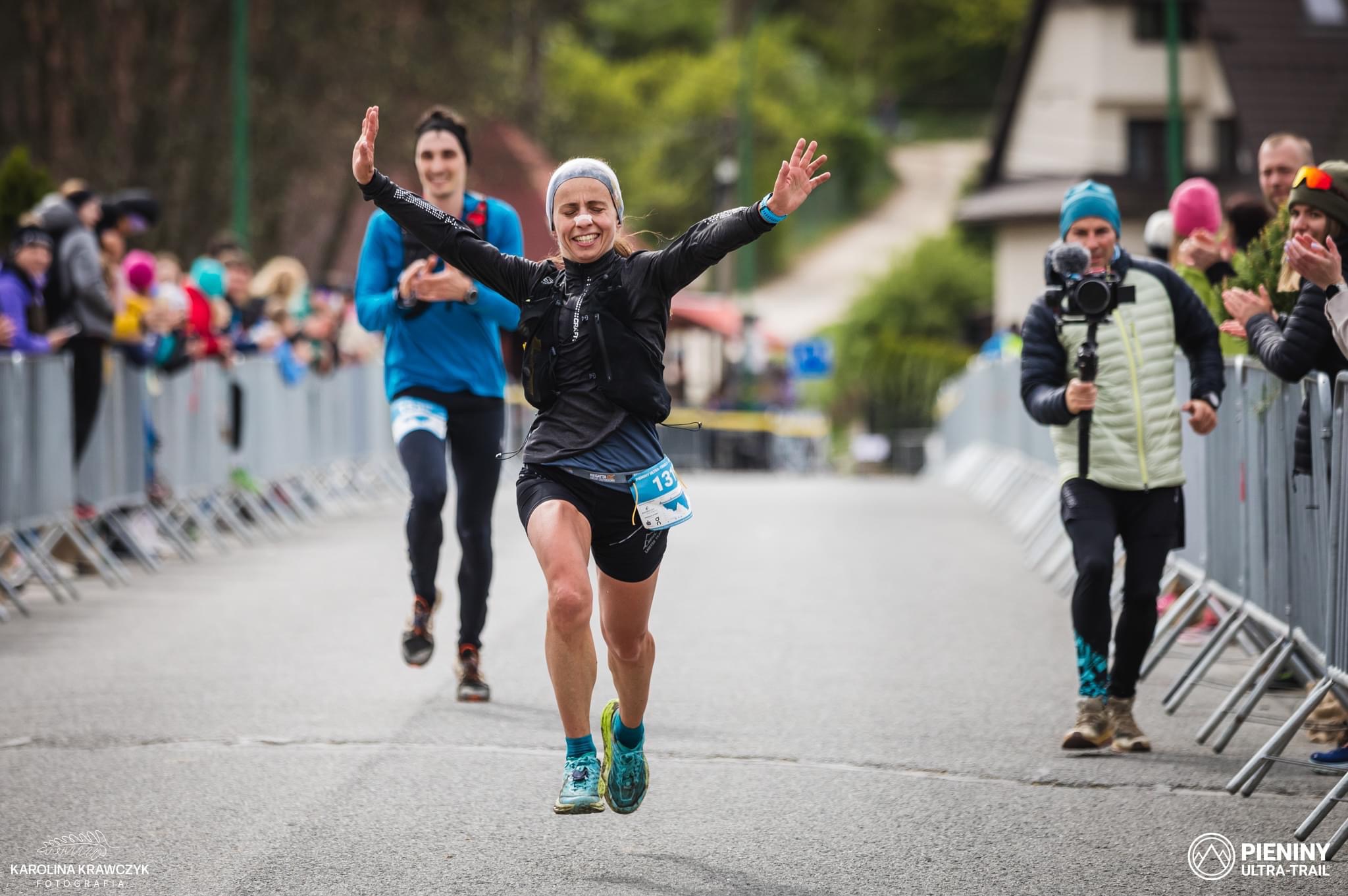 Uśmiechnięta biegaczka z numerem 131 na mecie biegu ulicznego, w tle kibice i kamerzysta.