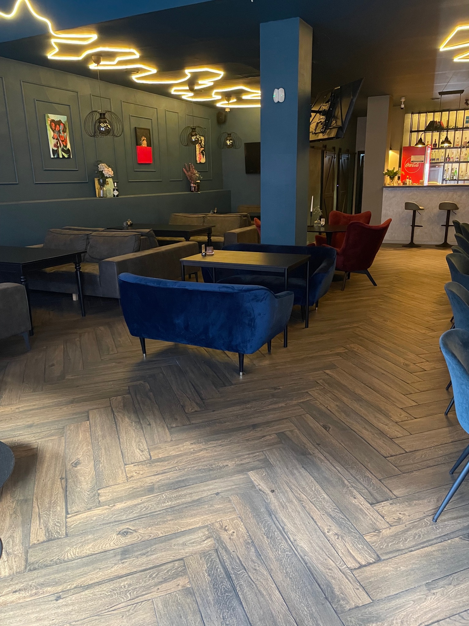 Wnętrze kawiarni z drewnianą podłogą w jodełkę, ciemnymi ścianami zdobionymi obrazami, nowoczesnymi lampami i wygodnymi sofami w odcieniach szarości i granatu, oraz barem w tle.