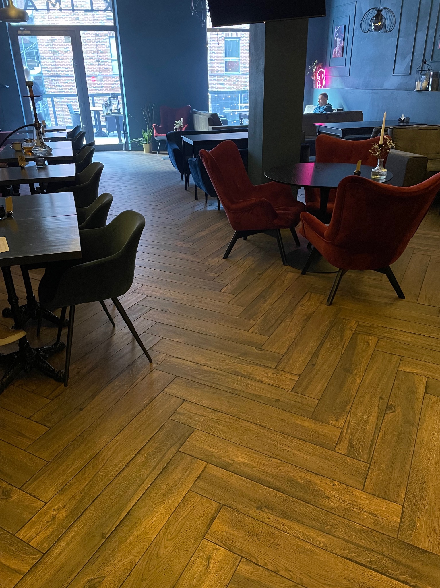 Wnętrze lokalu w Gdańsku z ciemnymi ścianami, parkietem ułożonym w jodełkę, stolikami i krzesłami w odcieniach czerwieni i zieleni. W tle widoczna osoba siedząca na kanapie.