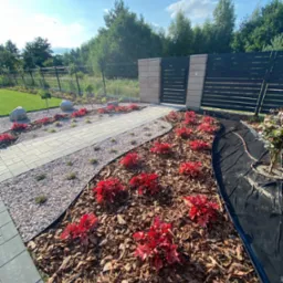 Ogród w Kałęczynie z ścieżką z kostki, żwirem i rabatami z kory oraz czerwonymi roślinami ozdobnymi, w tle nowoczesne ogrodzenie panelowe i brama.