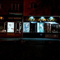Pracownia Plastyczna ZAH A.Dominiak - Nocny widok na fasadę sklepu z zegarkami 'Bajerski' z podświetlanym szyldem i plakatami reklamowymi w oknach wystawowych, obok sklep medyczny.