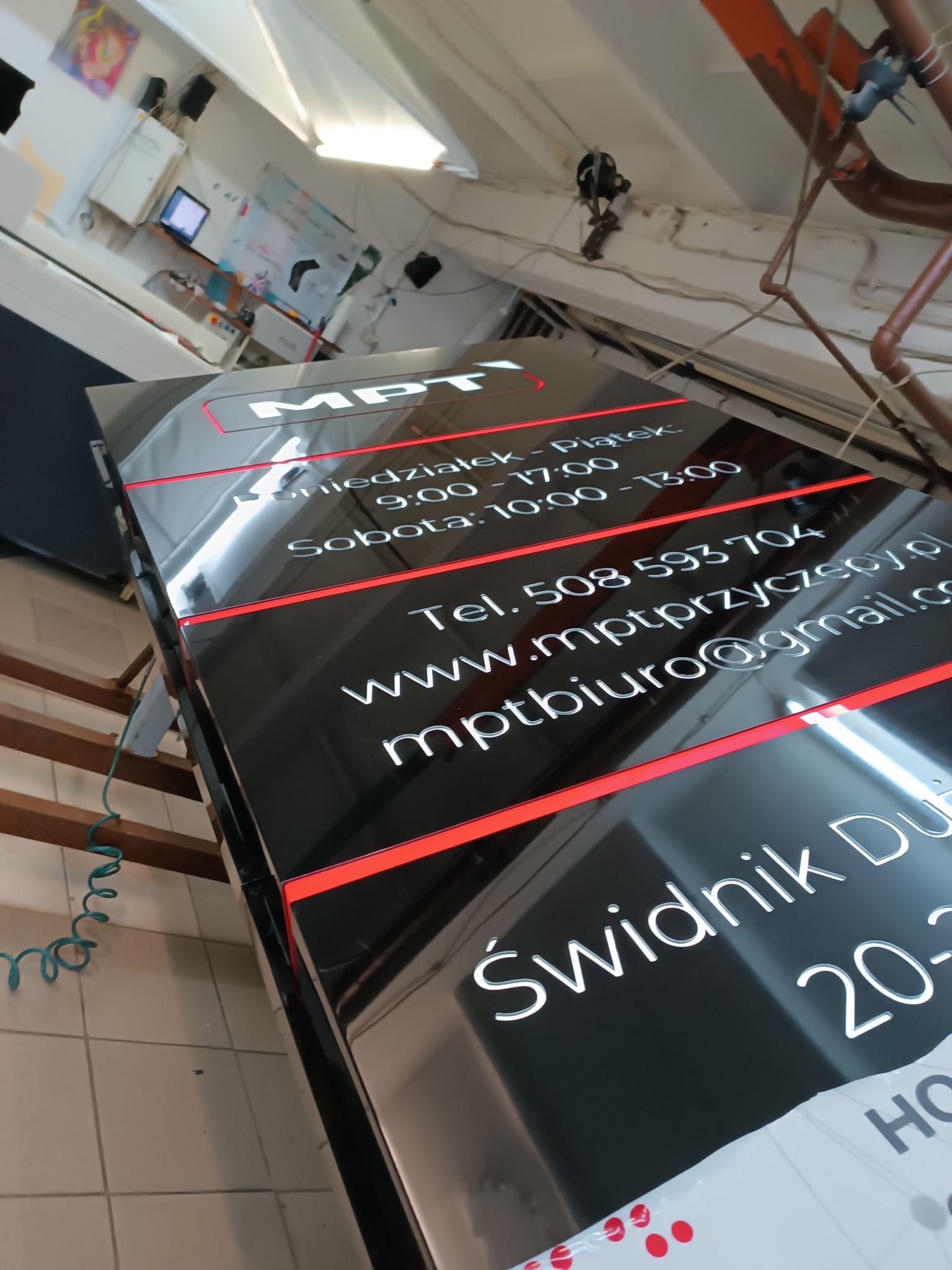 Czarna tablica informacyjna z białym napisem MPT, godzinami otwarcia, numerem telefonu, adresem strony internetowej i adresem e-mail, z czerwonymi liniami dekoracyjnymi, w tle fragment warsztatu.