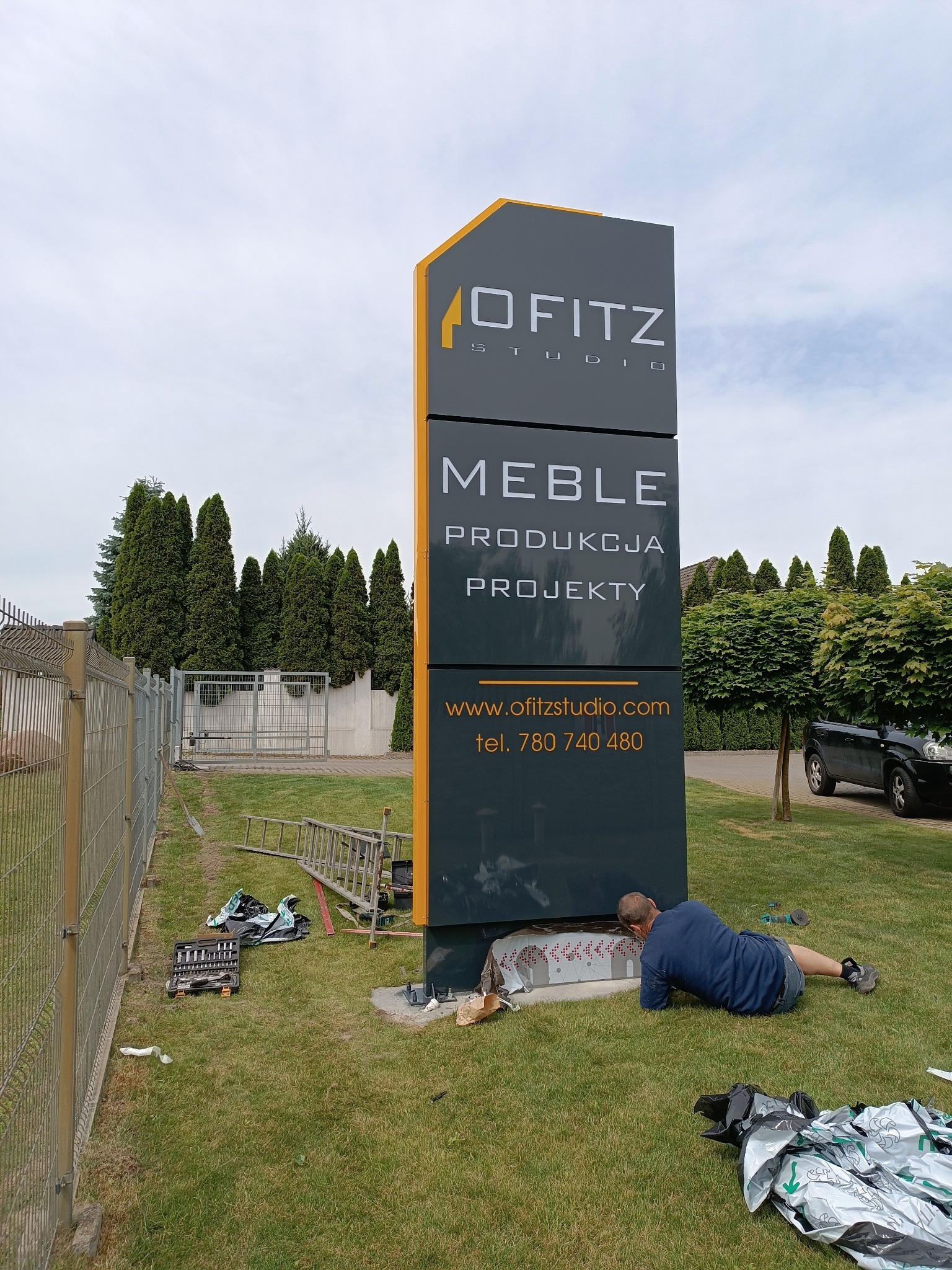 Montaż pionowego szyldu reklamowego firmy meblarskiej na trawniku, widoczny pracownik w trakcie instalacji, narzędzia i opakowania leżące obok.