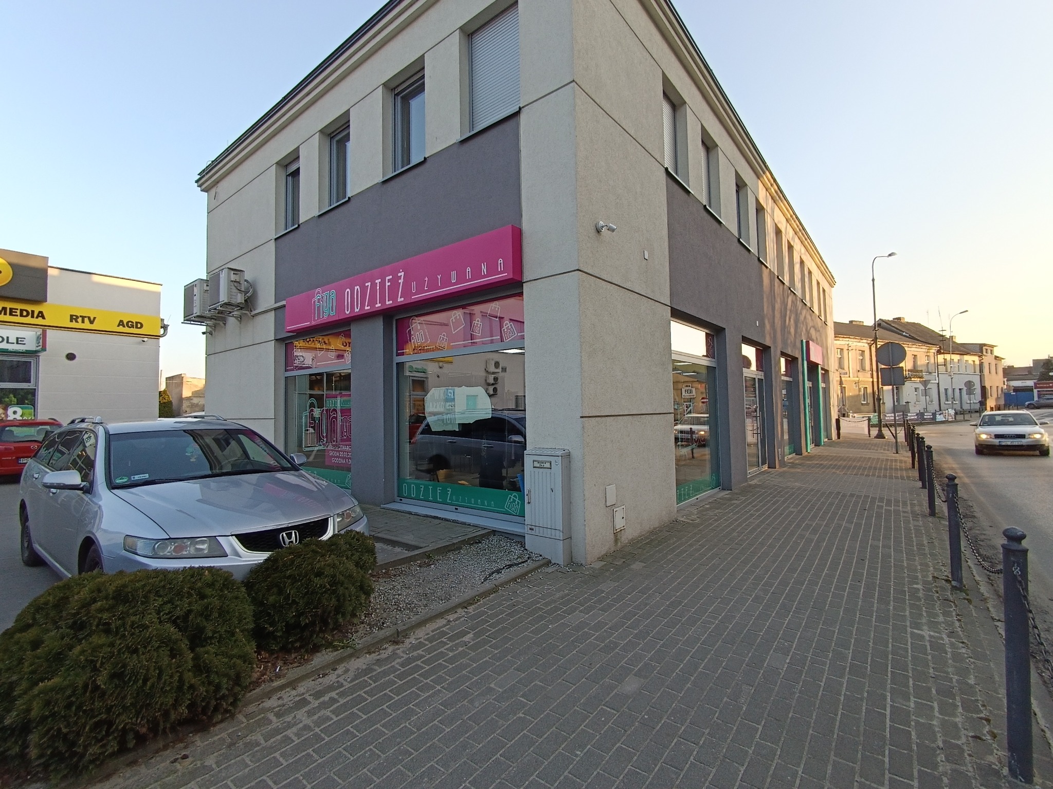 Dwupiętrowy budynek z szarymi i beżowymi ścianami, z szyldem sklepu z odzieżą używaną w kolorze różowym, widoczny z poziomu chodnika z kostki brukowej, z zaparkowanym srebrnym samochodem osobowym.
