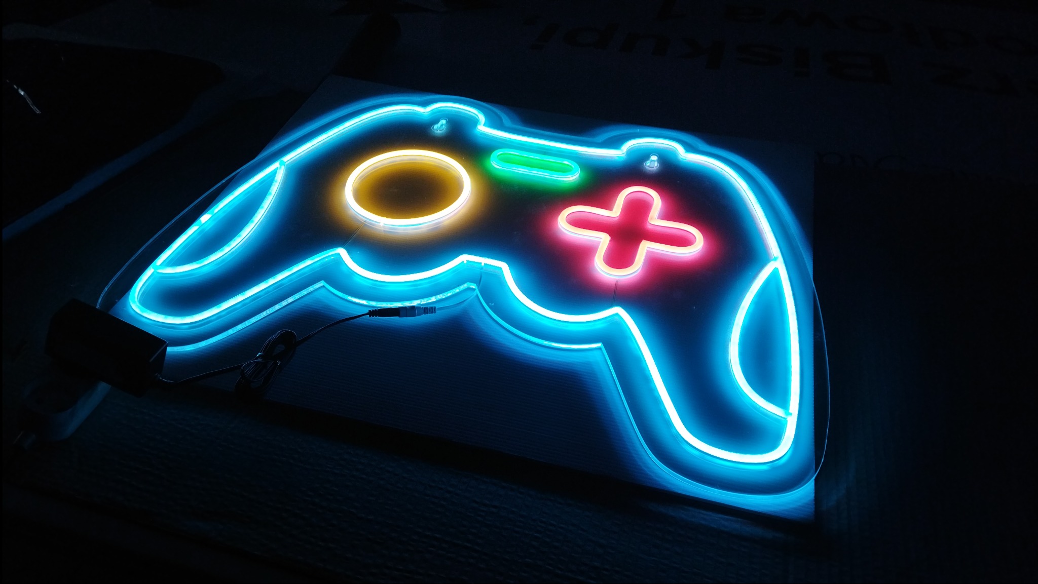 Niebieski neon w kształcie kontrolera do gier z podświetlanymi elementami: czerwony krzyżak, żółty okrąg i zielony prostokąt.