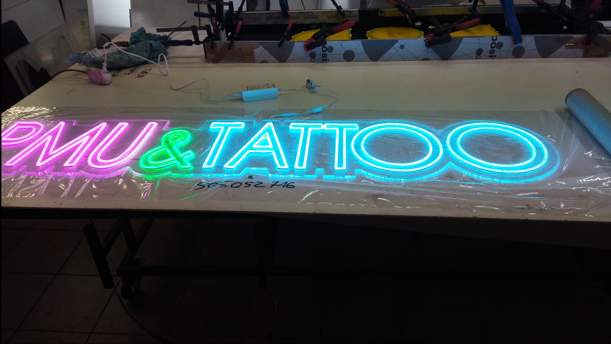 Napis neonowy PMU & TATTOO w kolorach różowym, zielonym i niebieskim, leżący na stole w warsztacie, widoczne przewody i zasilacz.