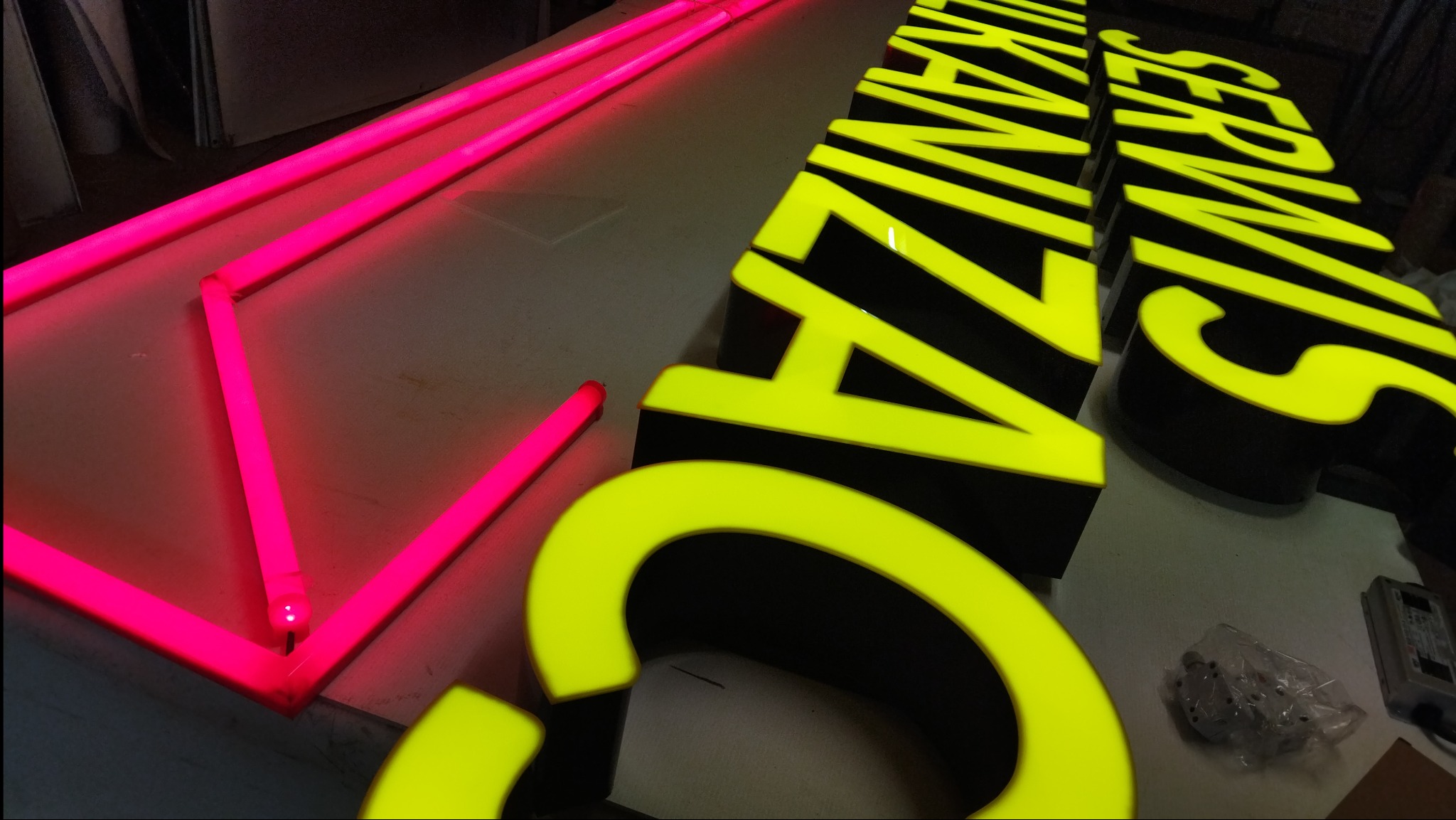 Neonowe litery z żółtym licem i czarnymi bokami, oraz różowe neonowe rurki w kształcie trójkątów, leżące na stole.