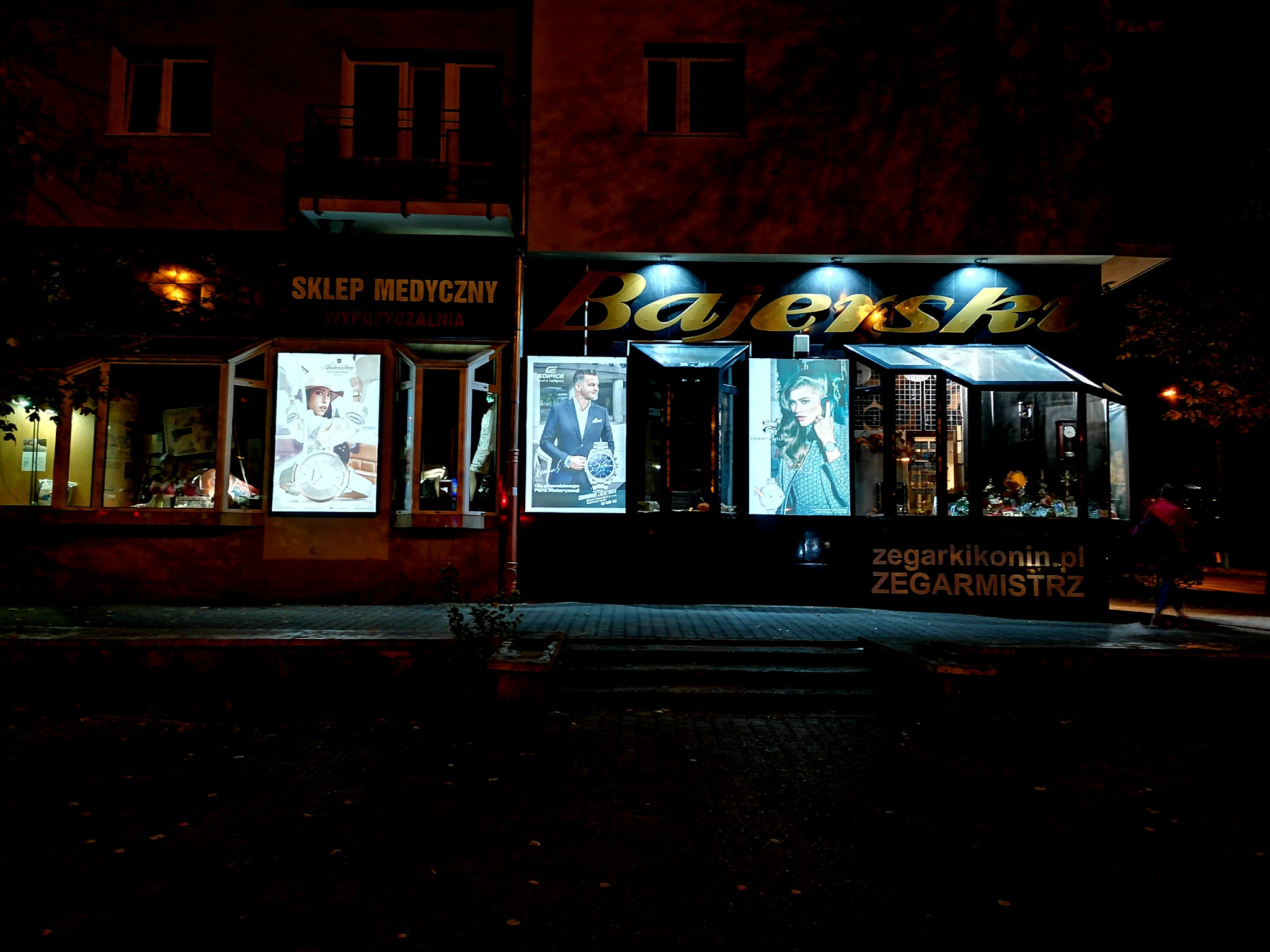 Nocny widok na fasadę sklepu z zegarkami 'Bajerski' z podświetlanym szyldem i plakatami reklamowymi w oknach wystawowych, obok sklep medyczny.