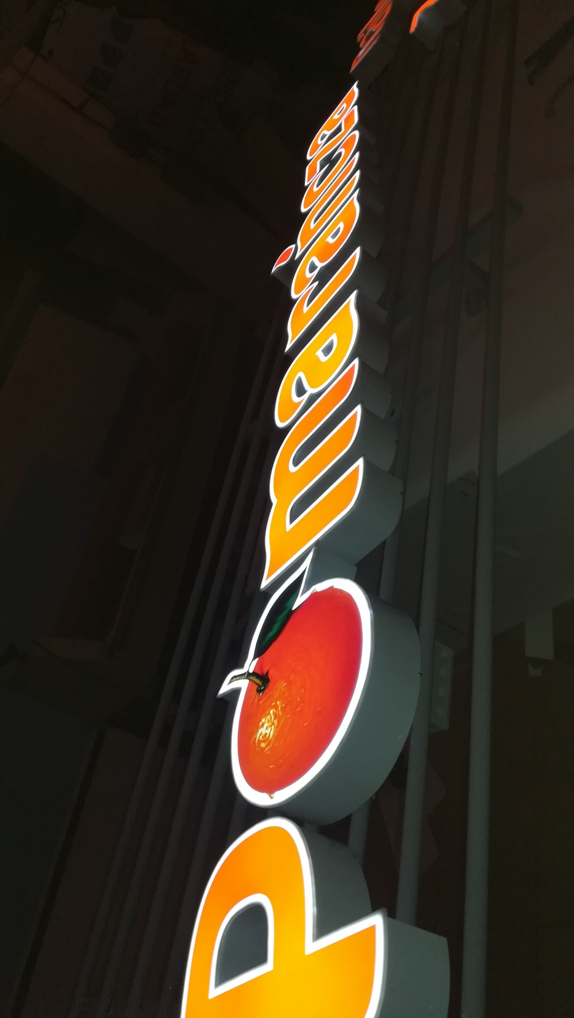 Pionowy, podświetlany szyld z nazwą firmy oraz elementem graficznym w kształcie pomarańczy z zielonym listkiem, widoczne z dołu pod kątem w ciemności.