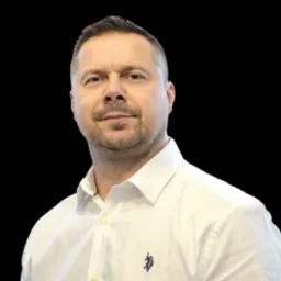 Portret mężczyzny w białej koszuli na czarnym tle, z delikatnym uśmiechem i widocznym logo na koszuli.