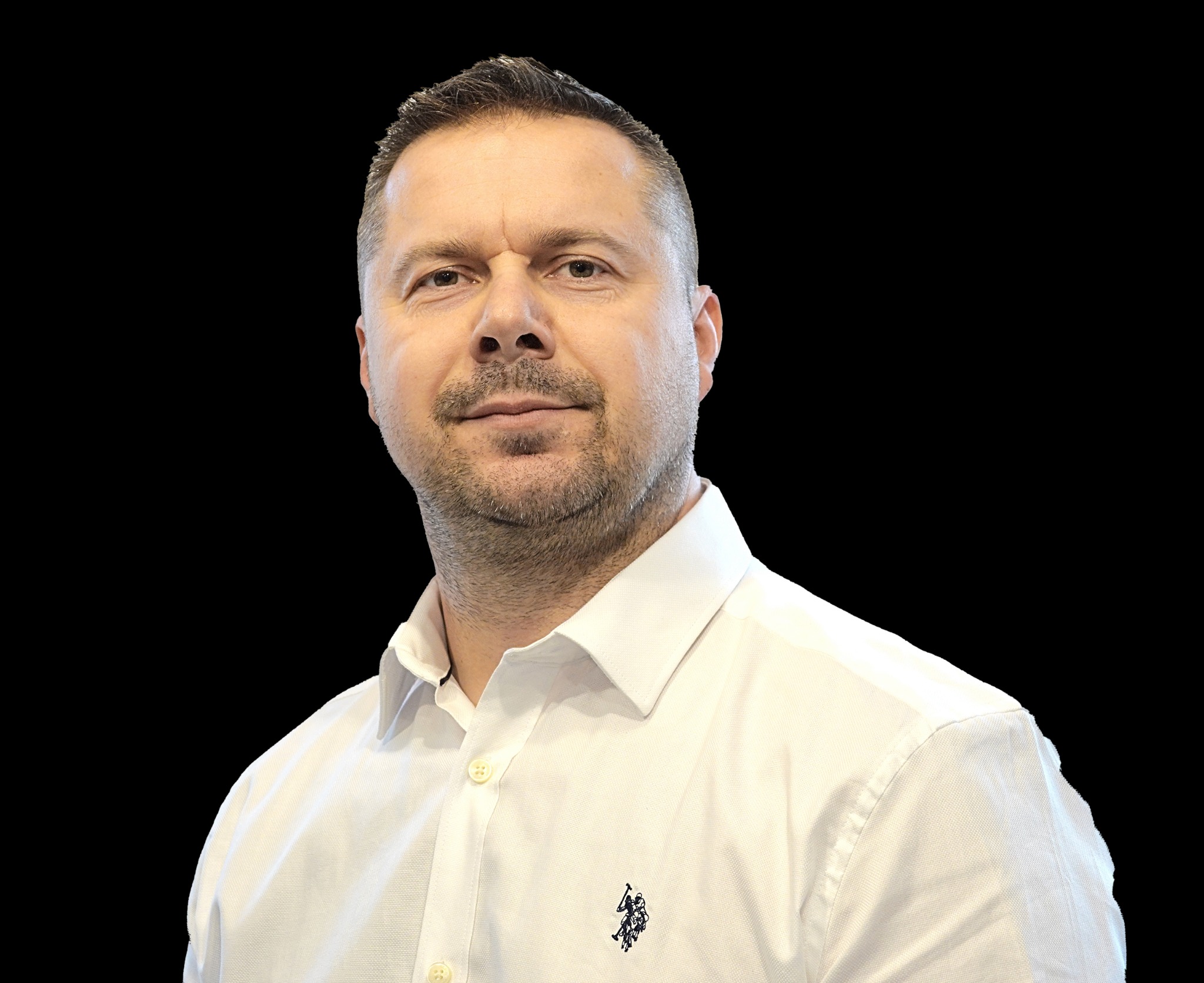 Portret mężczyzny w białej koszuli na czarnym tle, z delikatnym uśmiechem i widocznym logo na koszuli.