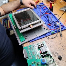 DSN-Elektronika - Technik w zegarku demontuje panel sterowania z zielonymi płytkami drukowanymi na warsztatowym stole z narzędziami i miernikami.