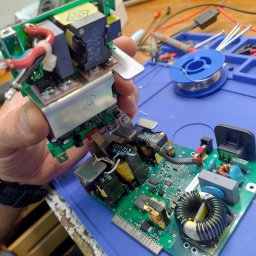 DSN-Elektronika - Serwis elektroniki: Ręka trzyma moduł elektroniczny z widocznymi kondensatorami, transformatorami i układami scalonymi na tle warsztatu z narzędziami i lutownicą.