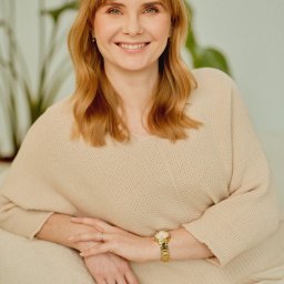 Gabinet Psychologiczny Magdalena Kaczmarek - Uśmiechnięta kobieta o rudoblond włosach w beżowym swetrze, siedząca na jasnej kanapie, z dłońmi splecionymi, złotym zegarkiem i pierścionkami. W tle rozmyta zieleń rośliny.