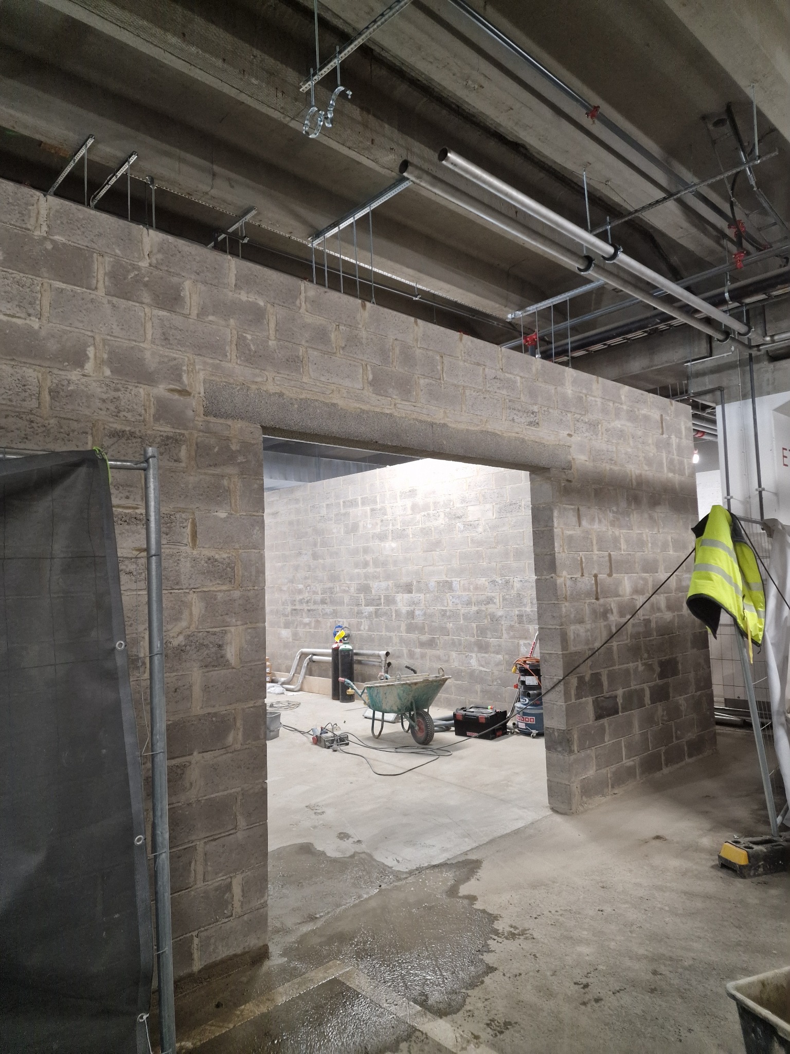 Wzniesiona ściana z szarego bloczka betonowego z otworem drzwiowym, widok na wnętrze z narzędziami i rurami pod sufitem. Żółta kamizelka odblaskowa w tle.