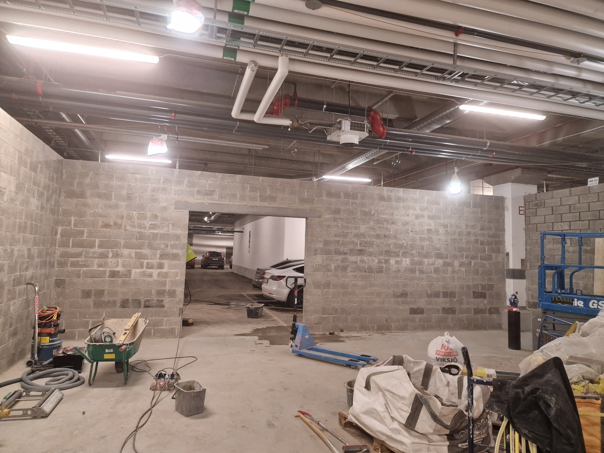 Budowa garażu podziemnego: surowe ściany z bloczków betonowych, narzędzia budowlane i samochody w tle. Widoczne instalacje pod sufitem.
