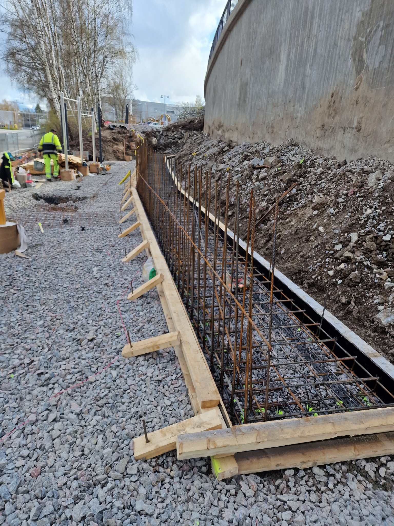 Zbrojenie i szalunek pod betonową konstrukcję oporową na żwirowym podłożu. Widoczny fragment betonowej ściany i pracownik w odblaskowym ubraniu w tle.