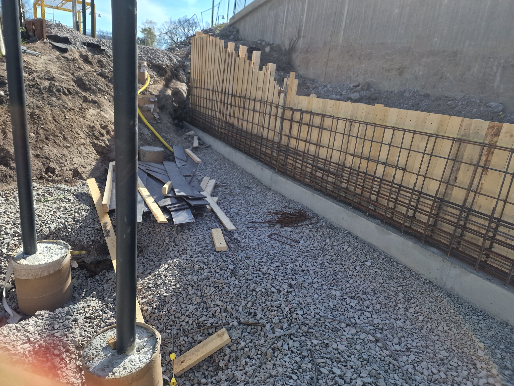 Zbrojenie ściany oporowej z drewnianym szalunkiem, widoczne elementy konstrukcyjne i podłoże z kruszywa. Prace ziemne w tle, słupy w betonie. Kątowe ujęcie.