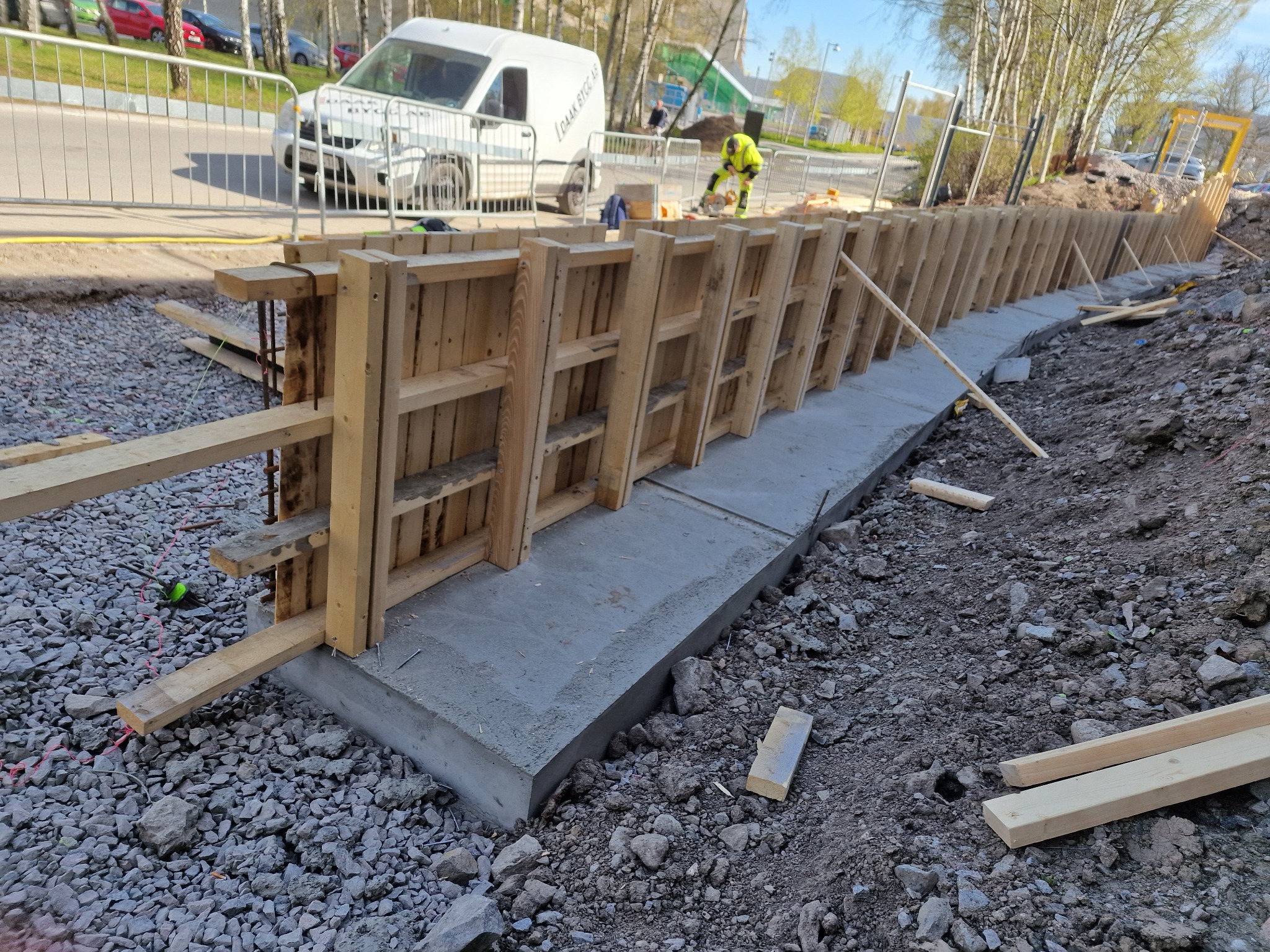 Betonowa podmurówka w drewnianym szalunku, widoczna zbrojenia. W tle pracownik w jaskrawym ubraniu i biały bus. Teren budowy.