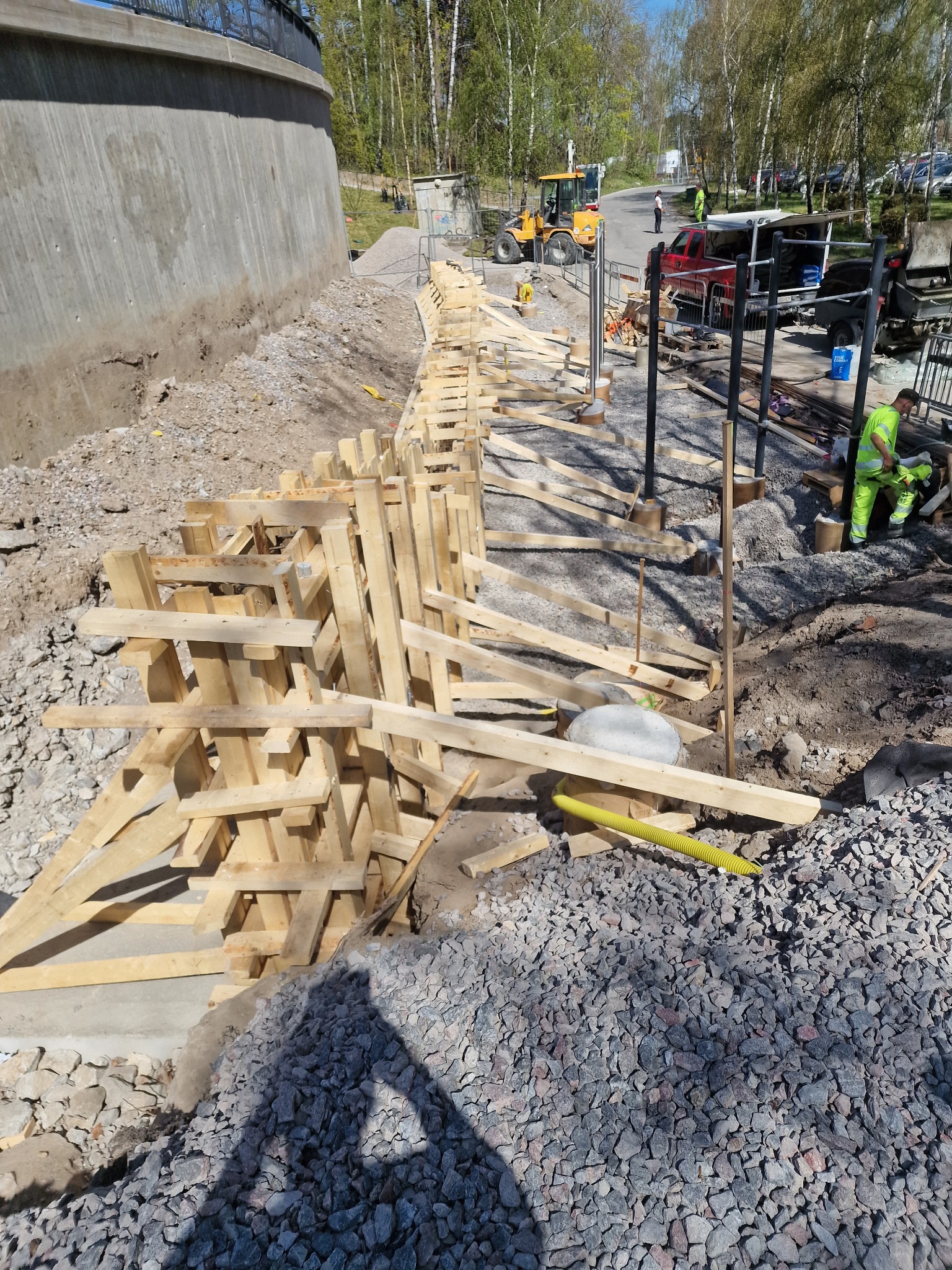Konstrukcja szalunkowa z drewna na placu budowy, widoczny betonowy mur oporowy, żwir i pracownik w odblaskowej odzieży. Prace przygotowawcze do betonowania.