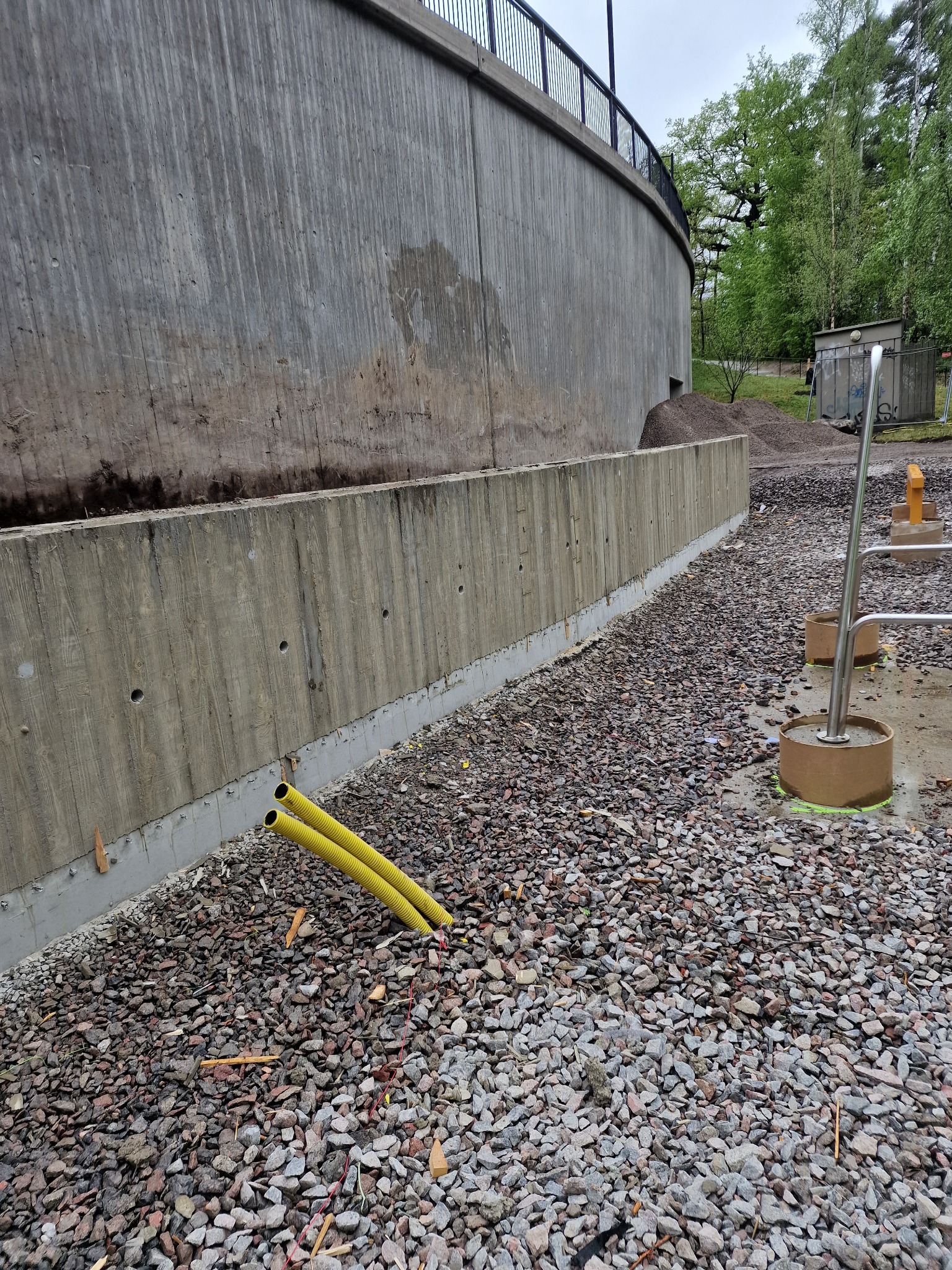 Surowy betonowy mur oporowy z żółtymi rurami w ziemi, otoczony żwirem. Widoczne ślady po szalunkach i niedoskonałości betonu, co dodaje industrialnego charakteru.