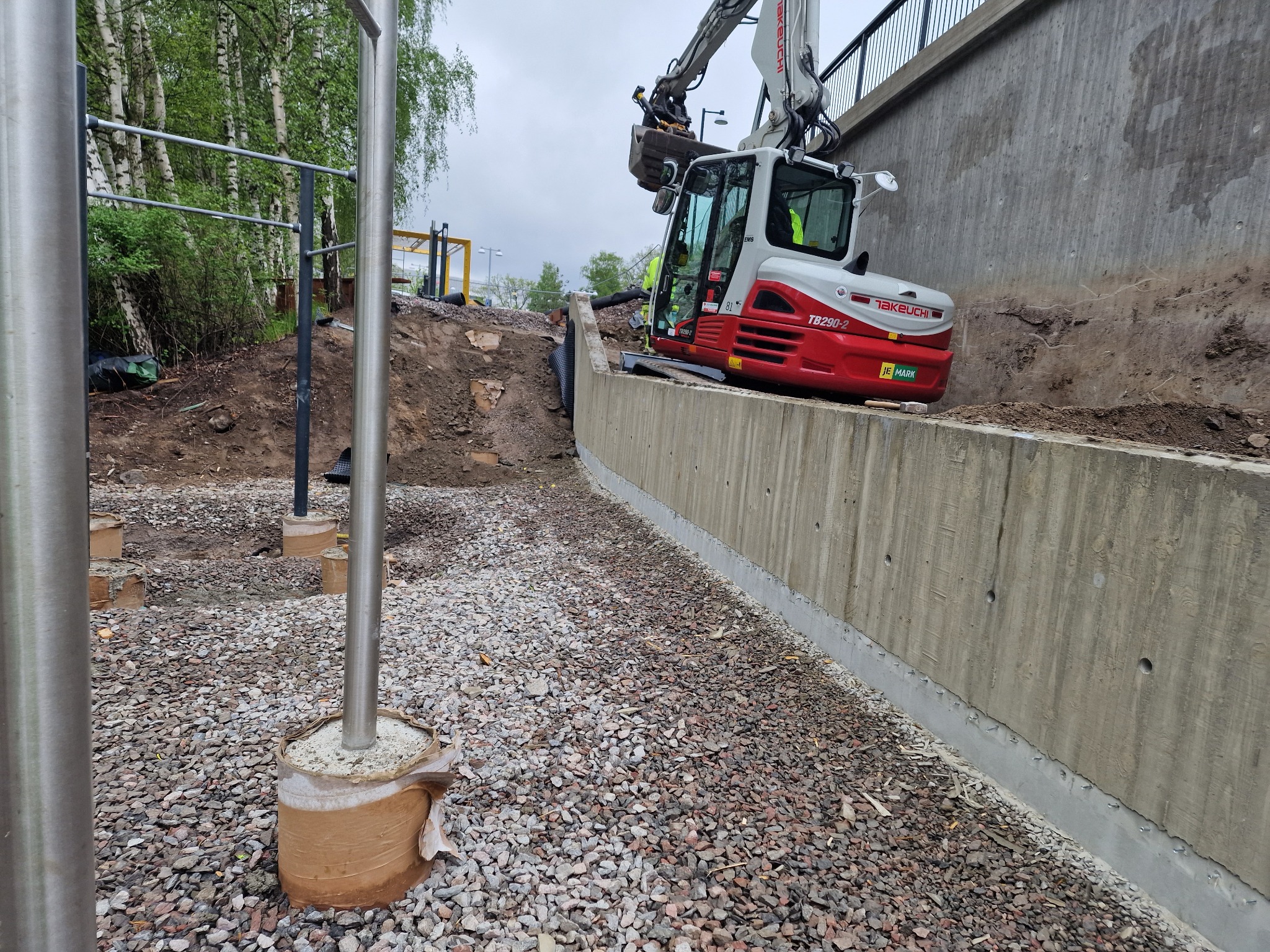 Prace ziemne przy betonowym murze oporowym, koparka Takeuchi TB290-2 w akcji. Widoczne przygotowania podłoża pod konstrukcję, teren wysypany drobnym kamieniem.