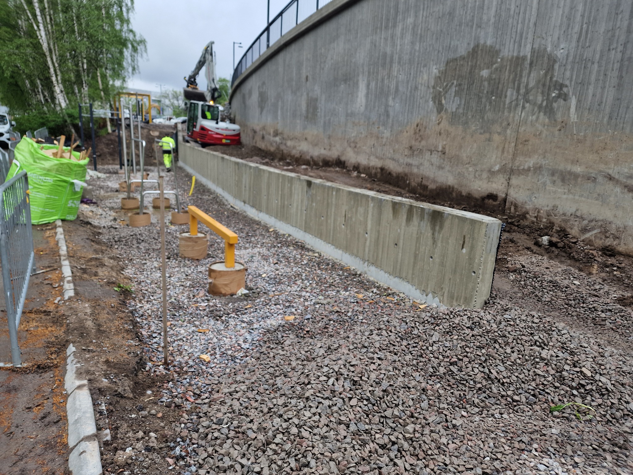 Budowa muru oporowego z betonu, podsypka z kamienia, w tle koparka i pracownik w odblaskowej kamizelce. Widok na plac budowy w pochmurny dzień.