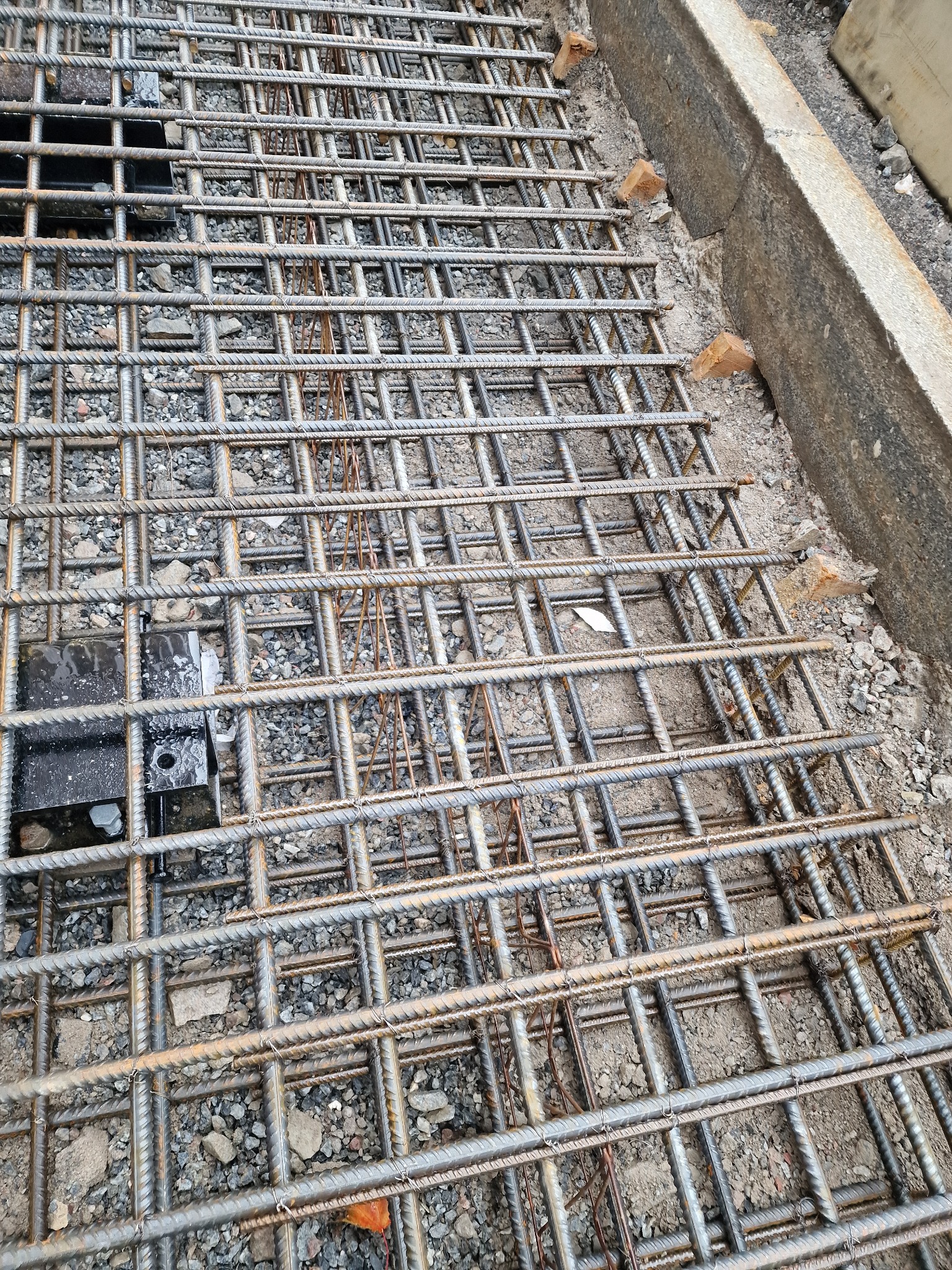 Zbrojenie fundamentu z siatki stalowej, przygotowane do zalania betonem. Widoczne elementy dystansowe i szalunek. Detal z bliska, wysoka perspektywa.