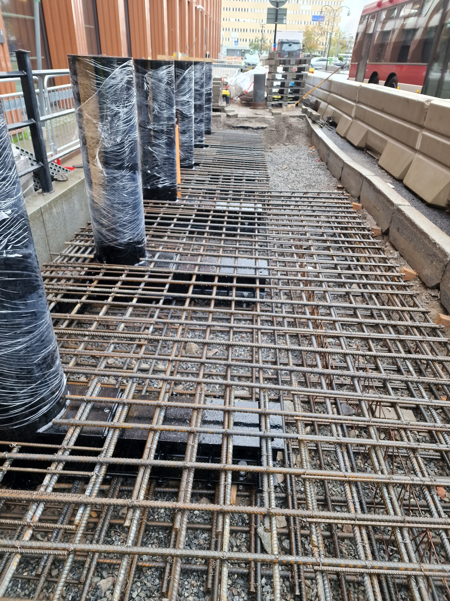 Zbrojenie terenu pod wylewkę, widoczne pionowe, czarne rury owinięte folią. W tle elementy infrastruktury miejskiej, w tym autobus.