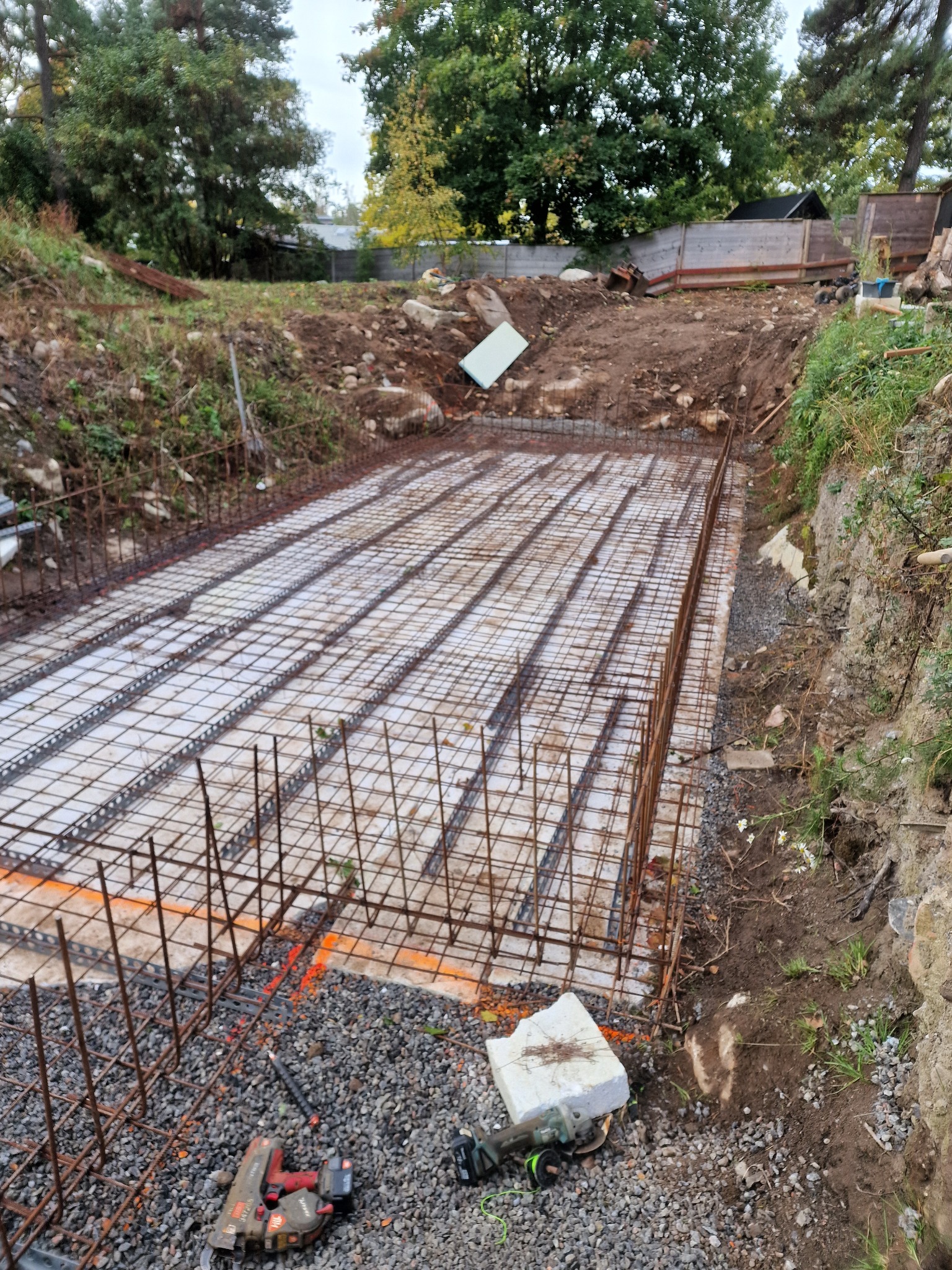 Zbrojenie fundamentów pod budynek, widoczne stalowe pręty ułożone na białej izolacji, narzędzia budowlane w tle, ziemia i roślinność dookoła.