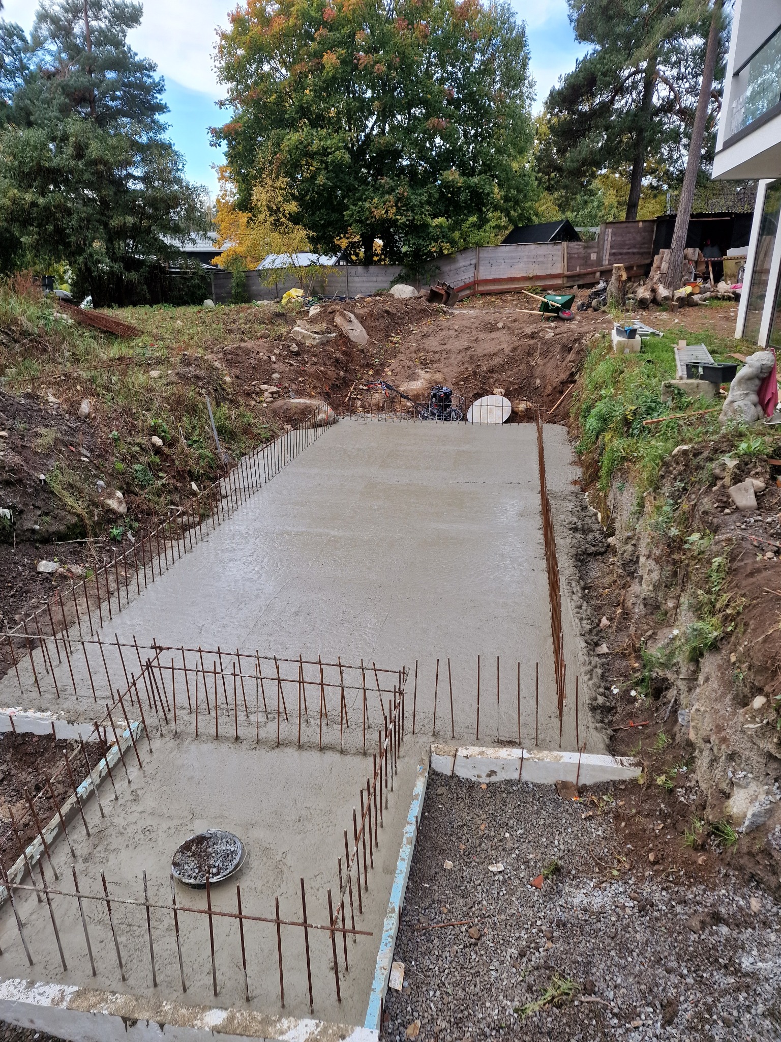 Wylany fundament zbrojony stalą na działce w wykopie. Widoczne szalunki i elementy instalacji. W tle drzewa i budynek w stylu nowoczesnym.