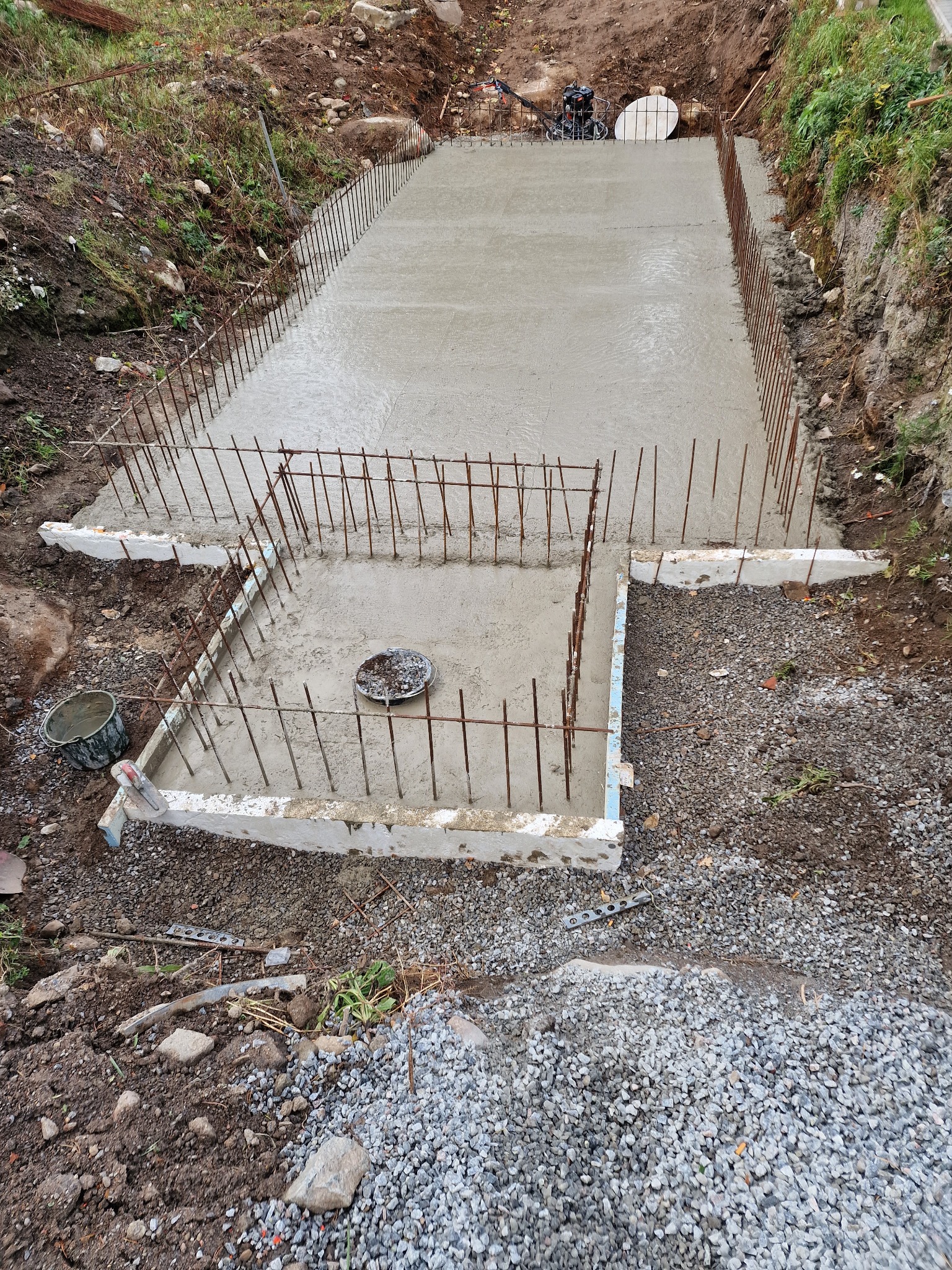 Wylany beton z wystającymi prętami zbrojeniowymi przygotowany pod fundament. Widoczne narzędzia i otoczenie placu budowy.