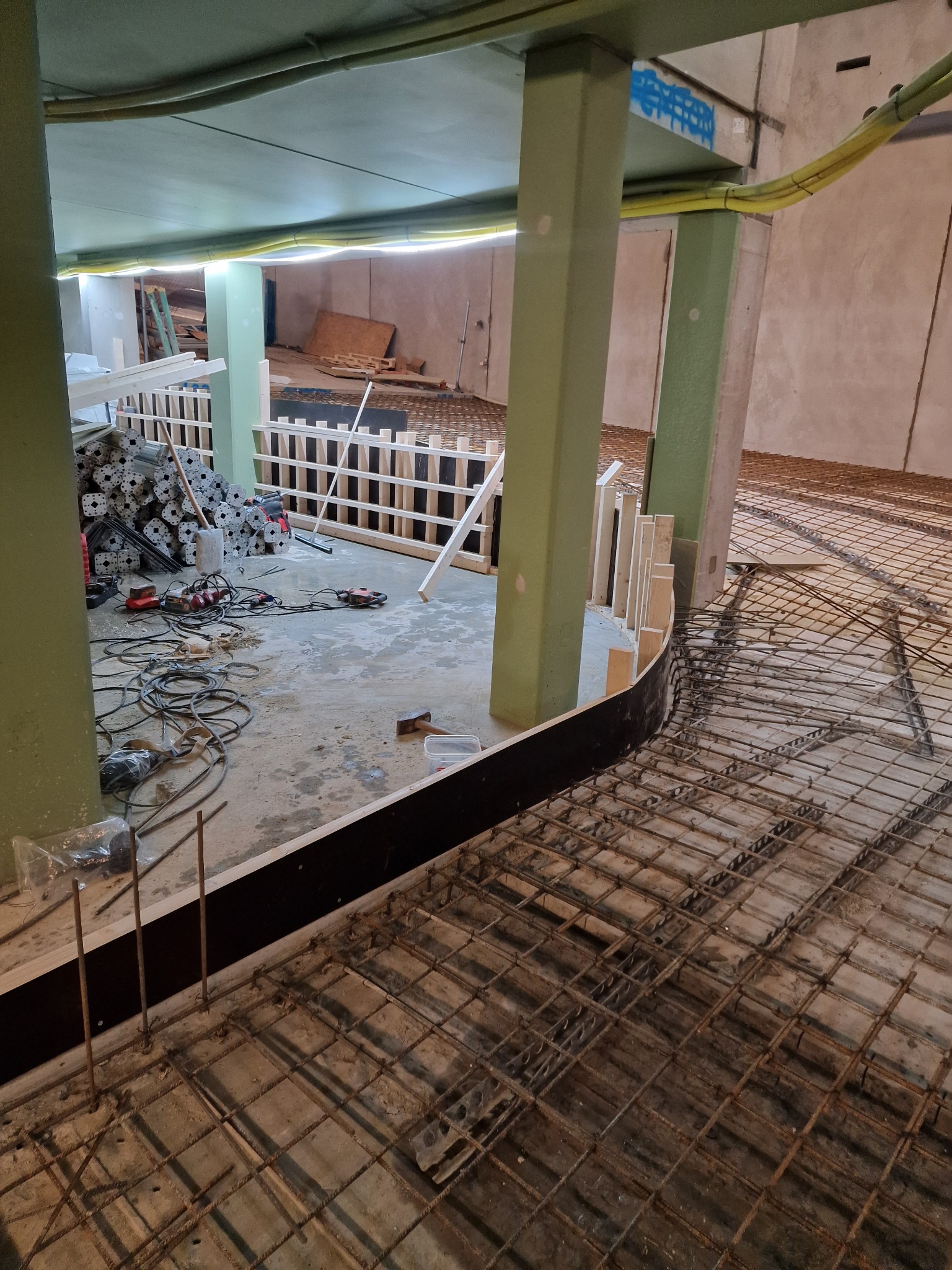 Zbrojenie podłoża z ułożonymi prętami i szalunkiem w kształcie łuku. Widoczne narzędzia, kable i podpory. Surowy stan prac wewnątrz budynku z kolumnami.