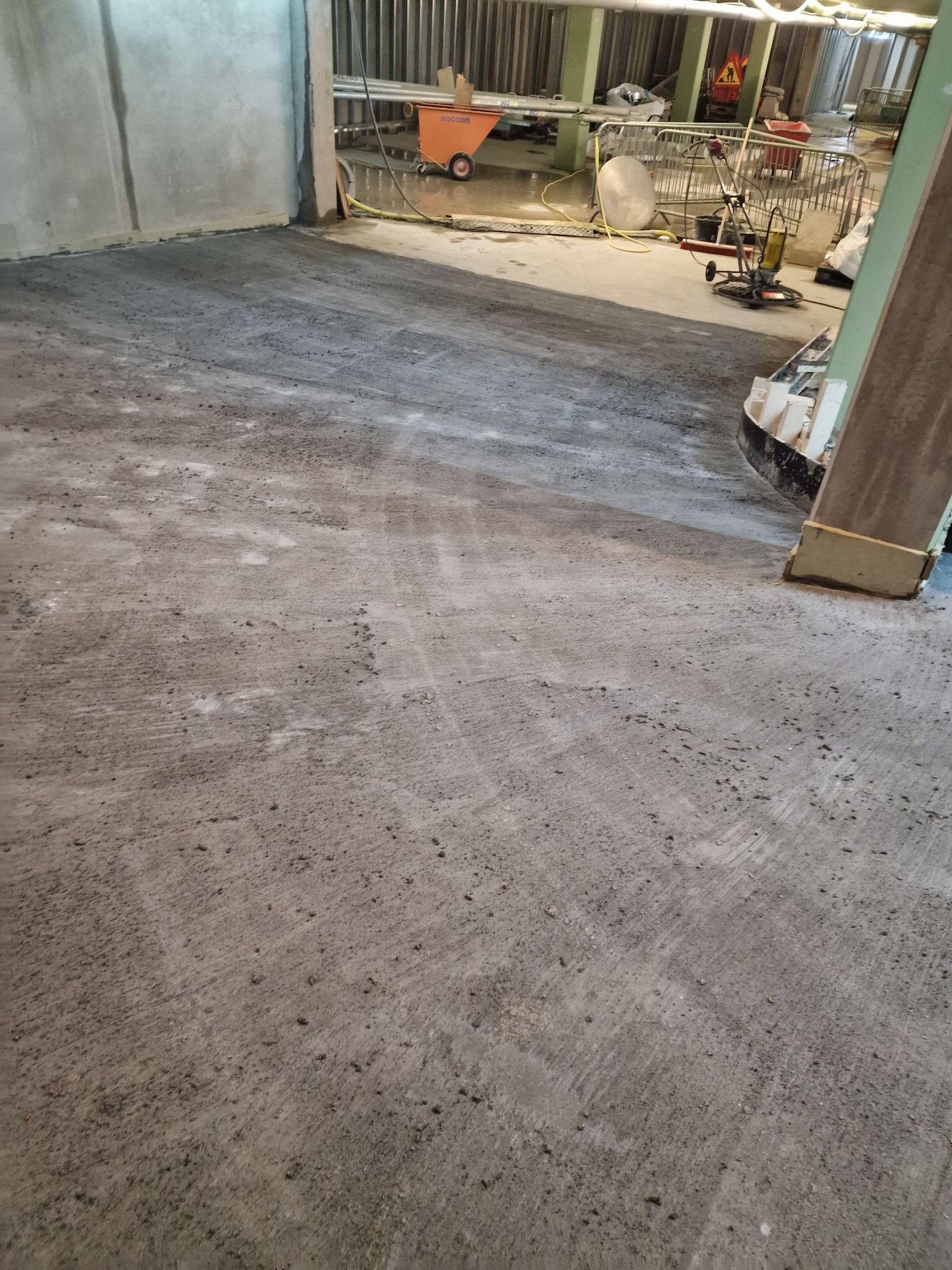 Wyrównana, szara betonowa podłoga w trakcie prac wykończeniowych, widoczne ślady po maszynie szlifującej, w tle narzędzia i materiały budowlane.