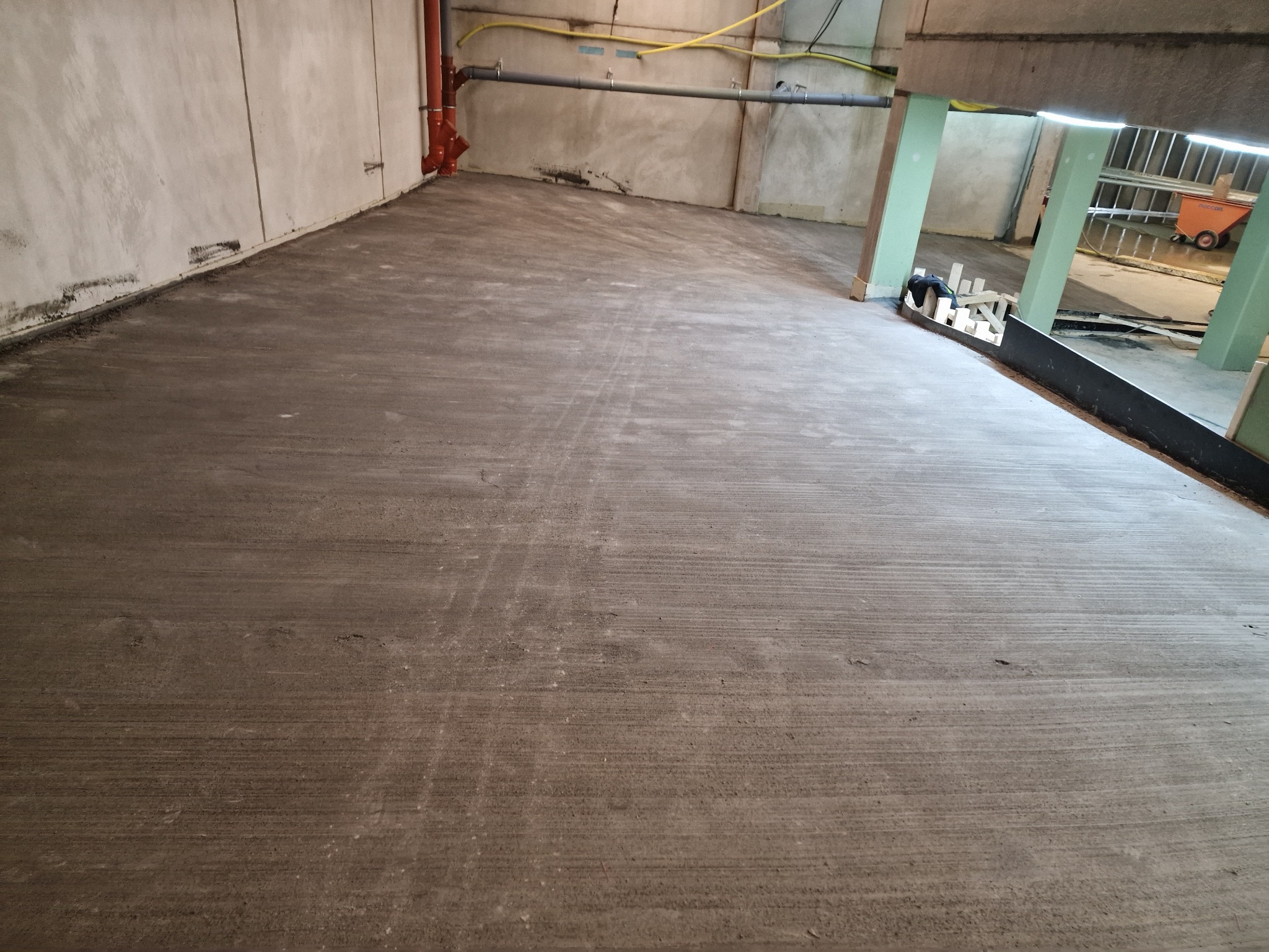 Wyrównana, szara wylewka podłogowa w surowym wnętrzu. Widoczne ślady po narzędziach, rury instalacyjne pod sufitem i fragment konstrukcji wsporczej.