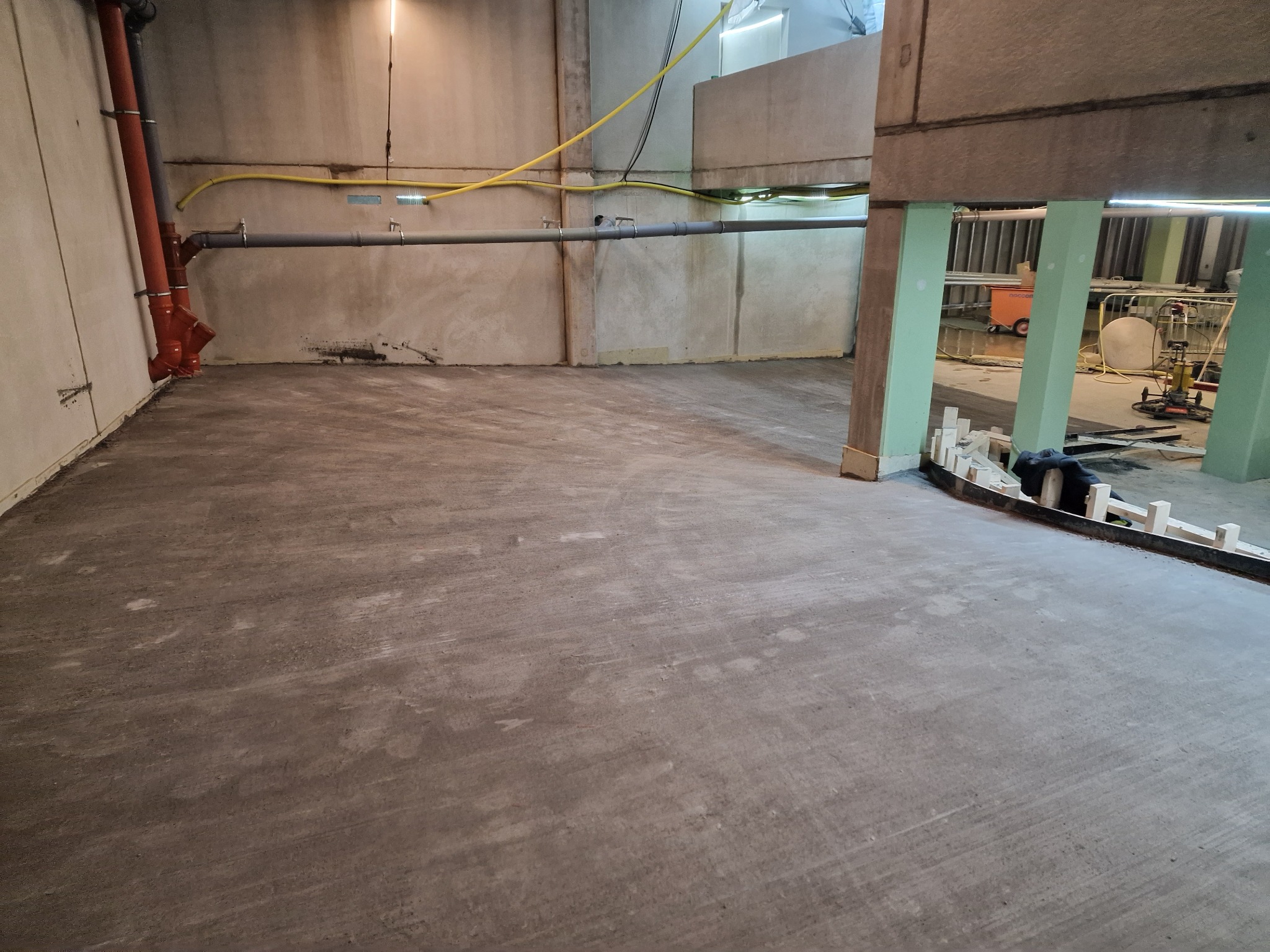 Wnętrze w trakcie prac wykończeniowych: surowa podłoga betonowa, widoczne instalacje pod sufitem i przygotowane szalunki. Widok na dalszą część pomieszczenia.