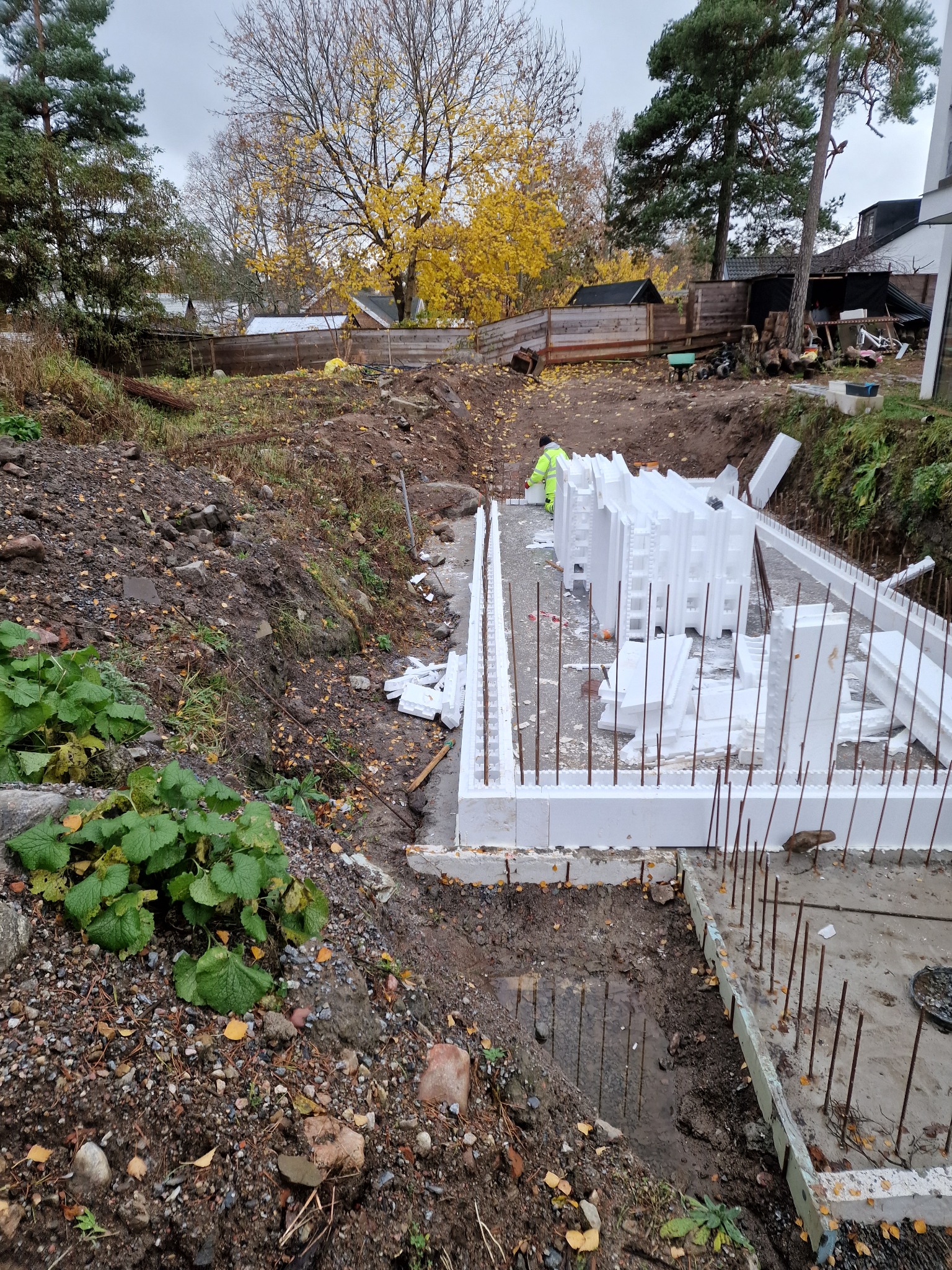 Budowa fundamentów domu z użyciem białych, styropianowych bloczków szalunkowych i zbrojenia stalowego. Widoczny wykop, ziemia i jesienne drzewa w tle.