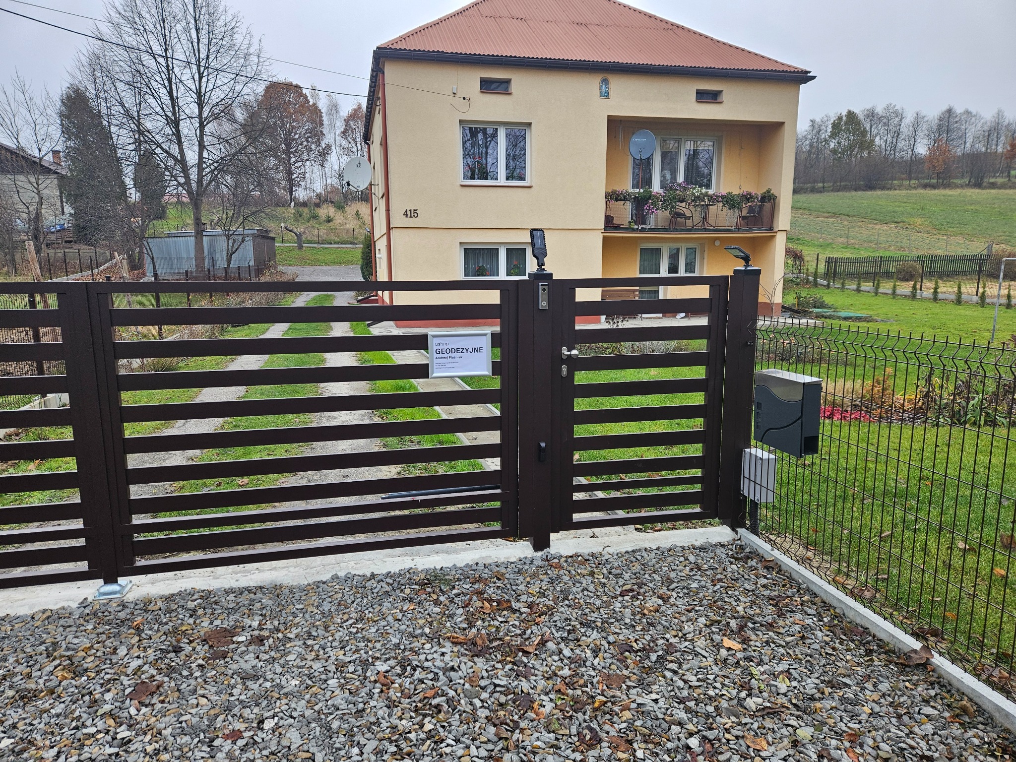 Nowoczesna, brązowa brama posesji z automatyką, widoczna z perspektywy żwirowego podjazdu, na tle dwupiętrowego domu z beżową elewacją i numerem 415. Tabliczka informacyjna 'Usługi Geodezyjne'...