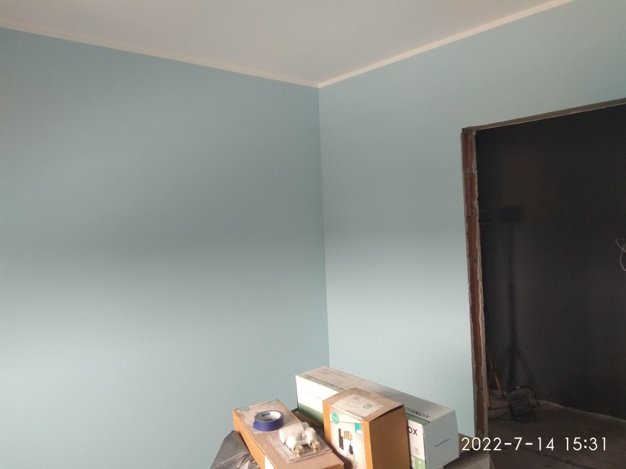 Świeżo pomalowane na błękitno ściany w rogu pokoju z widoczną białą listwą pod sufitem, z kartonami i akcesoriami do montażu lamp na pierwszym planie, oraz surowym otworem drzwiowym w tle.