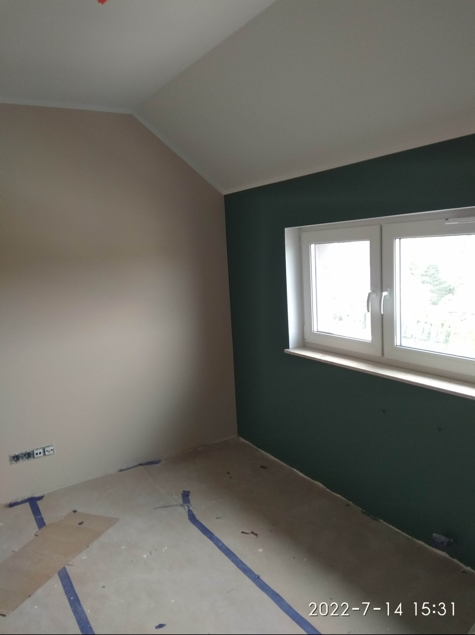 Pokój po częściowym pomalowaniu ścian: jedna ściana beżowa, druga ciemnozielona przy oknie, podłoga zabezpieczona taśmą malarską i kartonem, widoczne gniazdka elektryczne na ścianie.