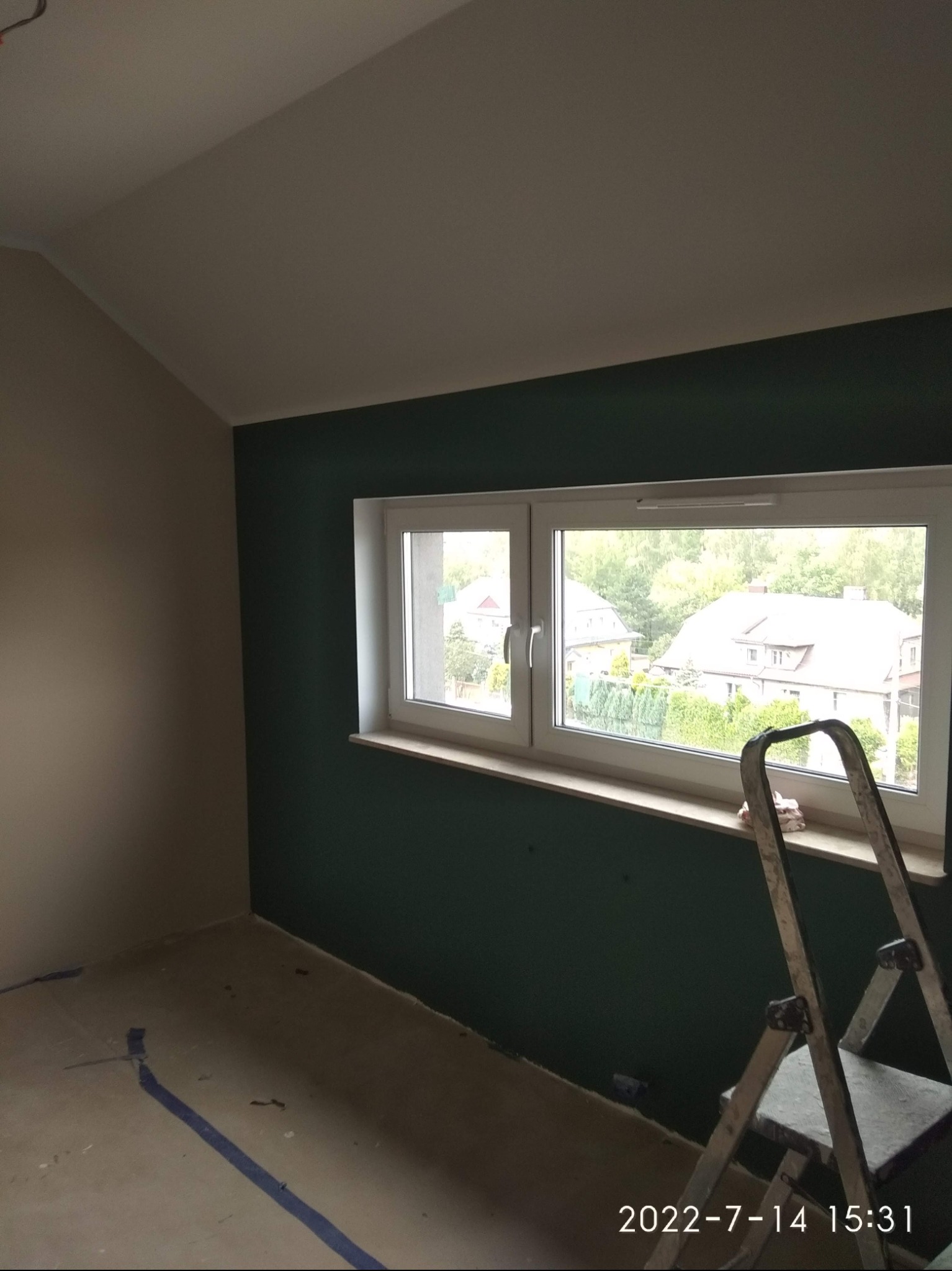 Pokój po częściowym pomalowaniu: jedna ściana w kolorze butelkowej zieleni, druga w jasnym beżu, okno z widokiem na domy i zieleń, drabina oparta o parapet, zabezpieczona podłoga.