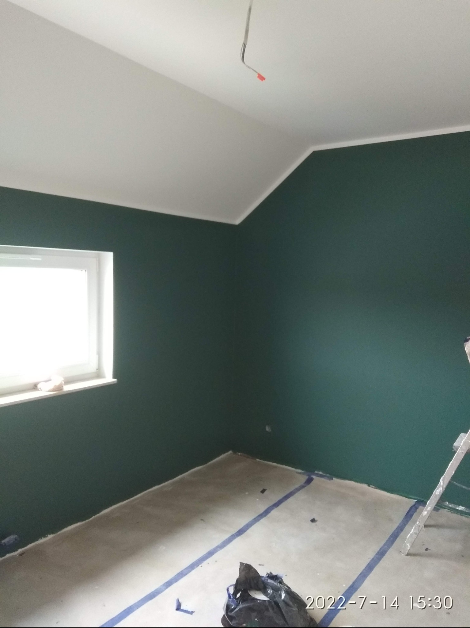 Pokój po malowaniu na ciemnozielony kolor, z białym sufitem i listwami, zabezpieczony niebieską taśmą malarską przy podłodze, widoczna drabina malarska.
