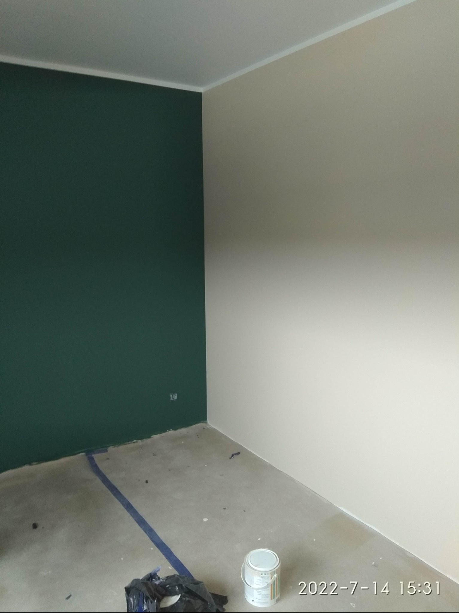 Pokój po częściowym pomalowaniu ścian na butelkową zieleń i beż, widoczna puszka farby i taśma malarska na podłodze.