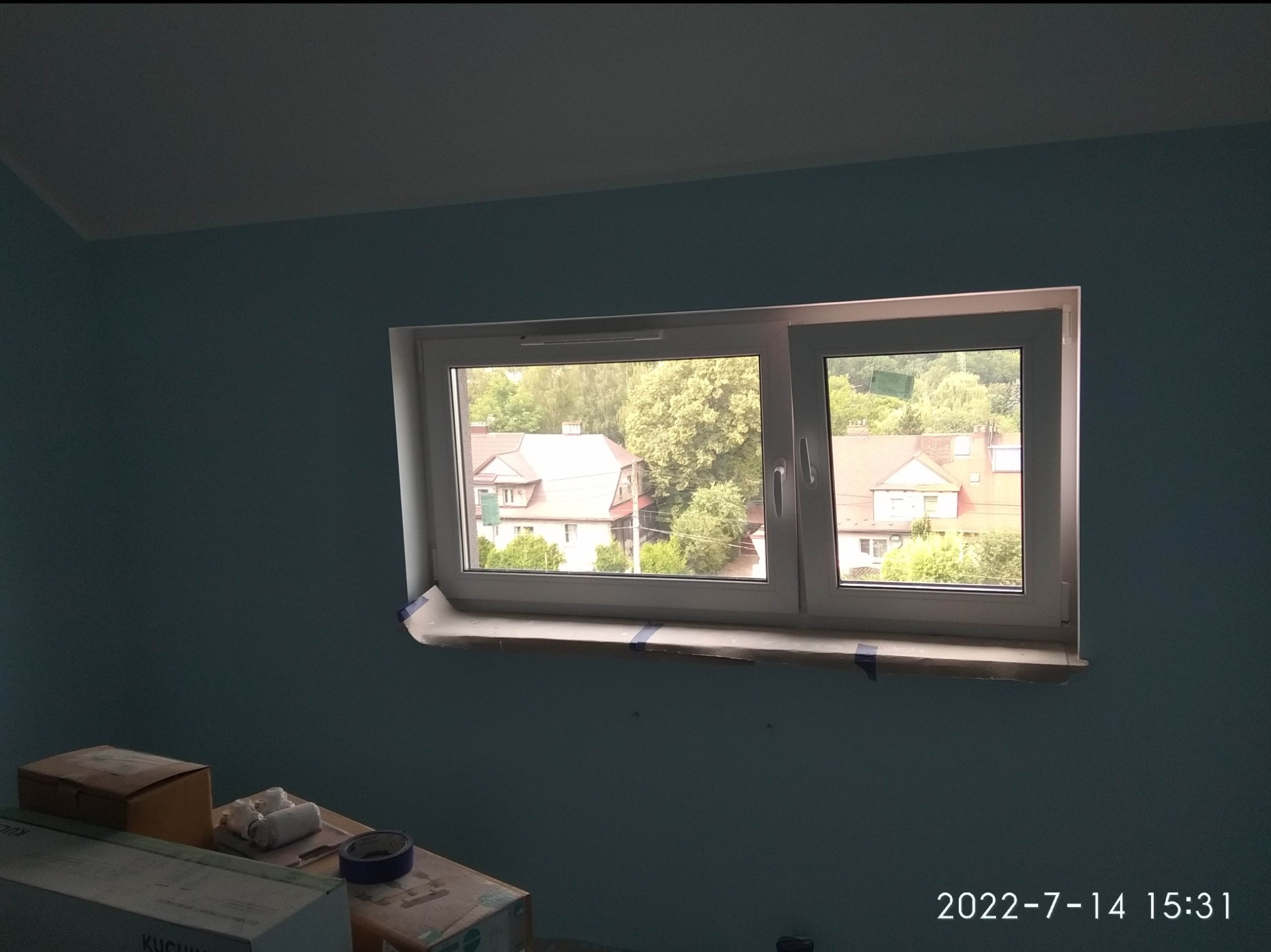 Niebieska ściana w trakcie malowania, okno zabezpieczone taśmą malarską i papierem, widoczne materiały malarskie w dolnym lewym rogu.